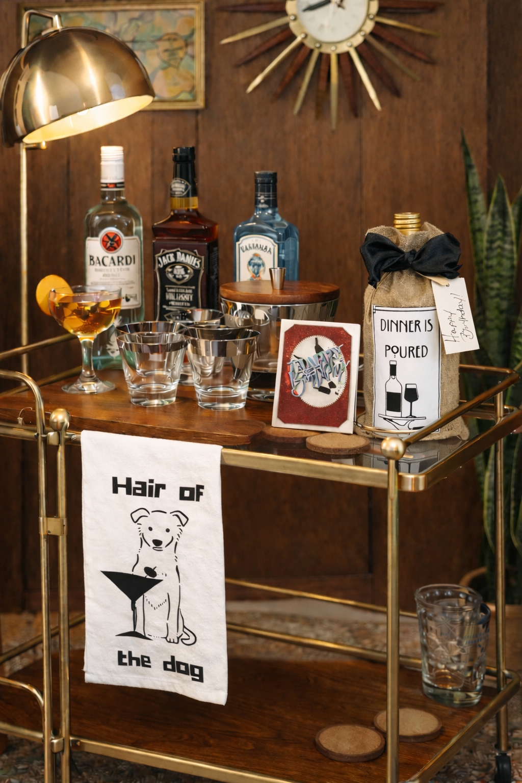 Midcentury modern bar cart styled with vintage cocktail glasses, liquor bottles, and brass accents, featuring a dog-themed “Hair of the Dog” cocktail tea towel, greeting card, and gift bag in a retro home bar setting.