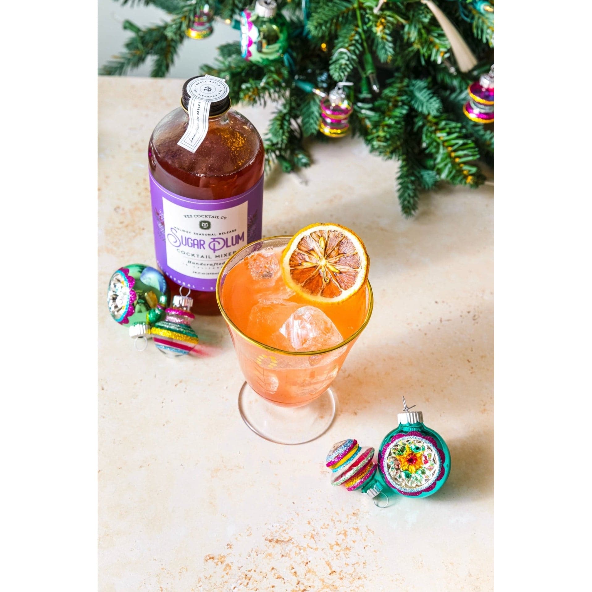 16 oz bottle of hand crafted Sugar Plum Cocktail Mixer made by Yes Cocktail Co displayed next to a cocktail made with the mix surrounded by Christmas ornaments and a tree in the background