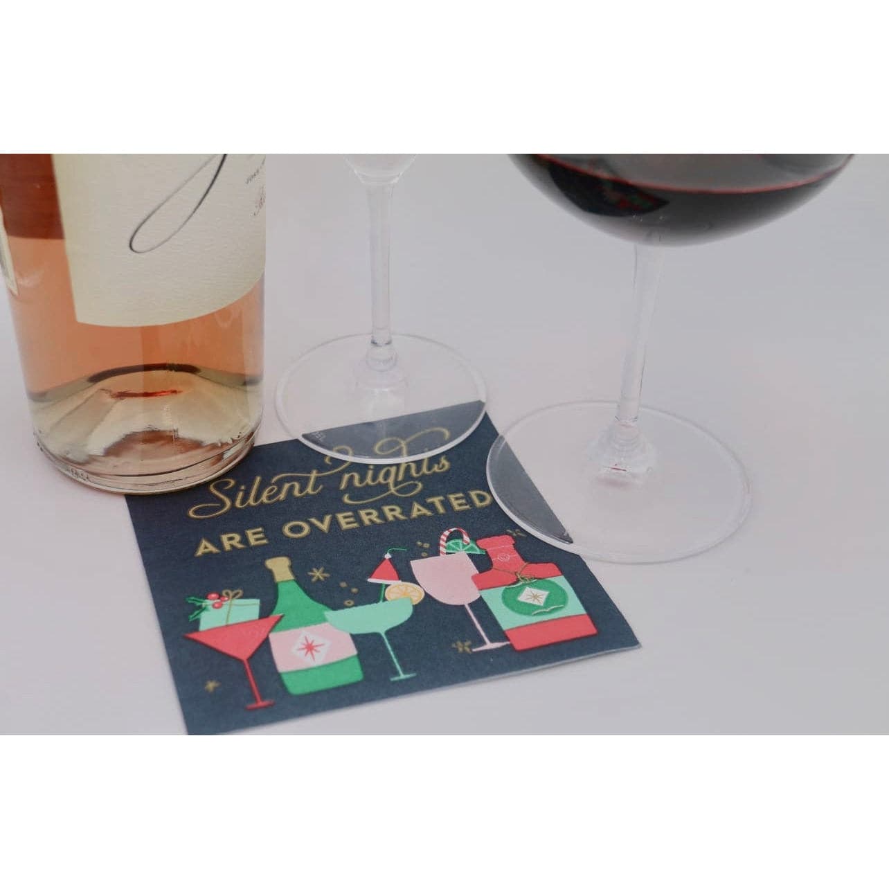 Christmas cocktail party napkins with a dark green background, differnt cocktail glassess and bottles are featured across the bottom in festive colors, and across the top in gold are the words "silent nights are overrated" displayed with a wine bottle and wine glasses