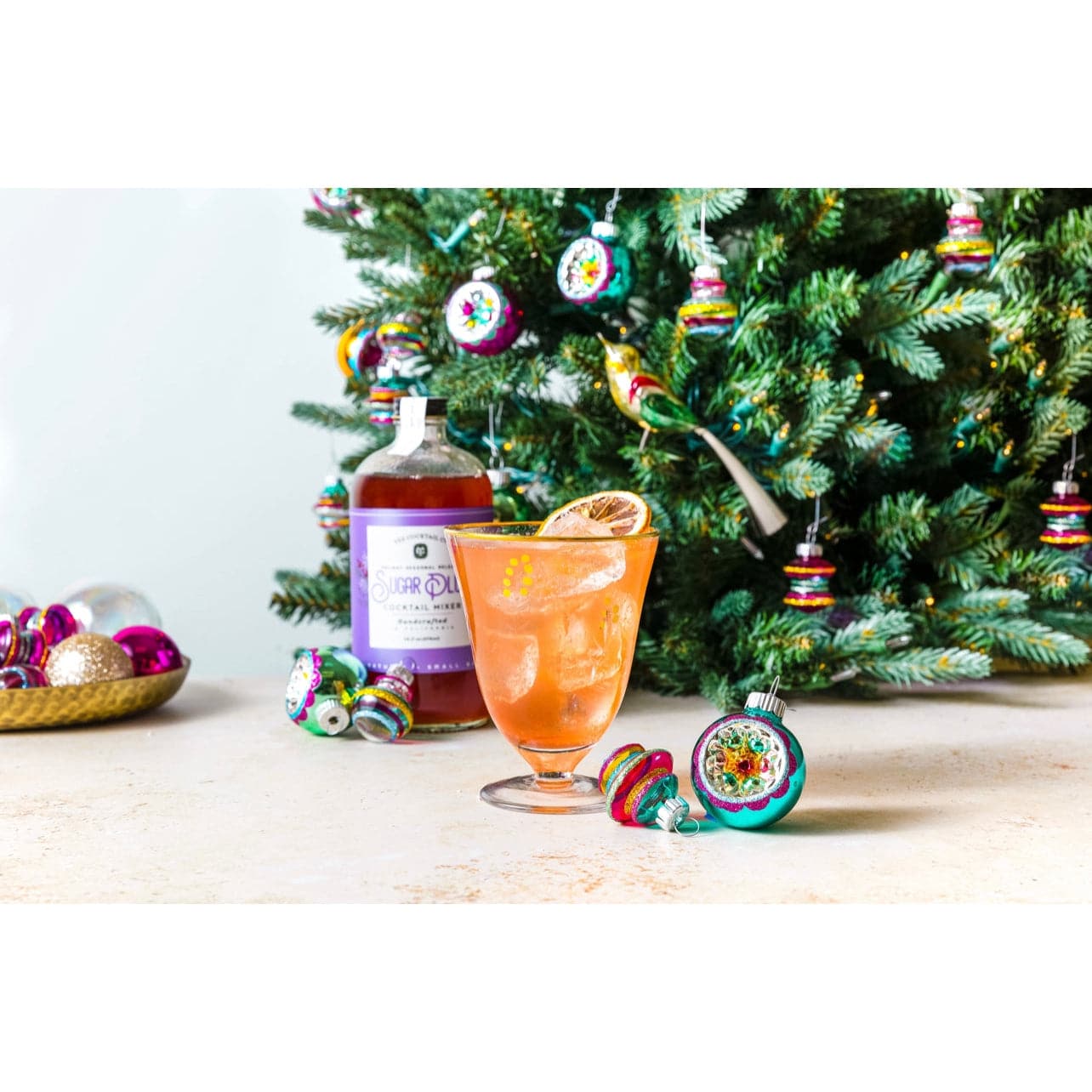 16 oz bottle of hand crafted Sugar Plum Cocktail Mixer made by Yes Cocktail Co displayed next to a cocktail made with the mix surrounded by Christmas ornaments and a tree in the background