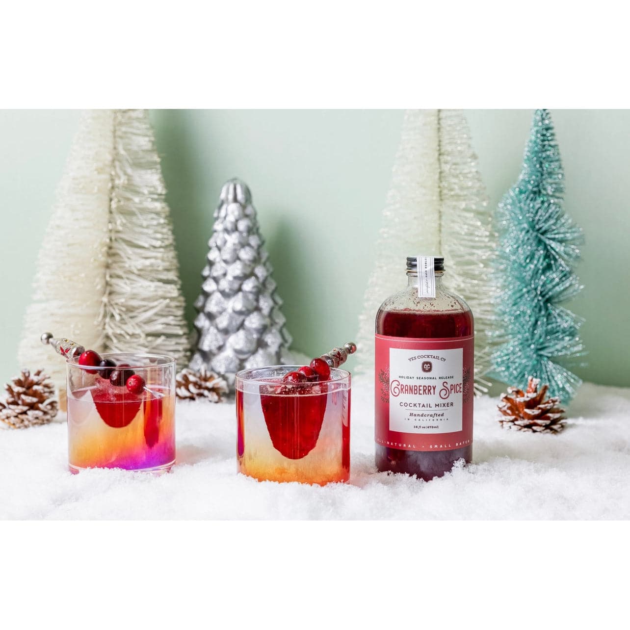 16 oz bottle of handcrafted Cranberry Spice Cocktail Mixer made by Yes Cocktail Co displayed with finished cocktails in iridescent double old fashioned glasses that are garnished with fresh cranberries. Fake snow is spread on the table with decorative Christmas tree shaped decorations in the background.