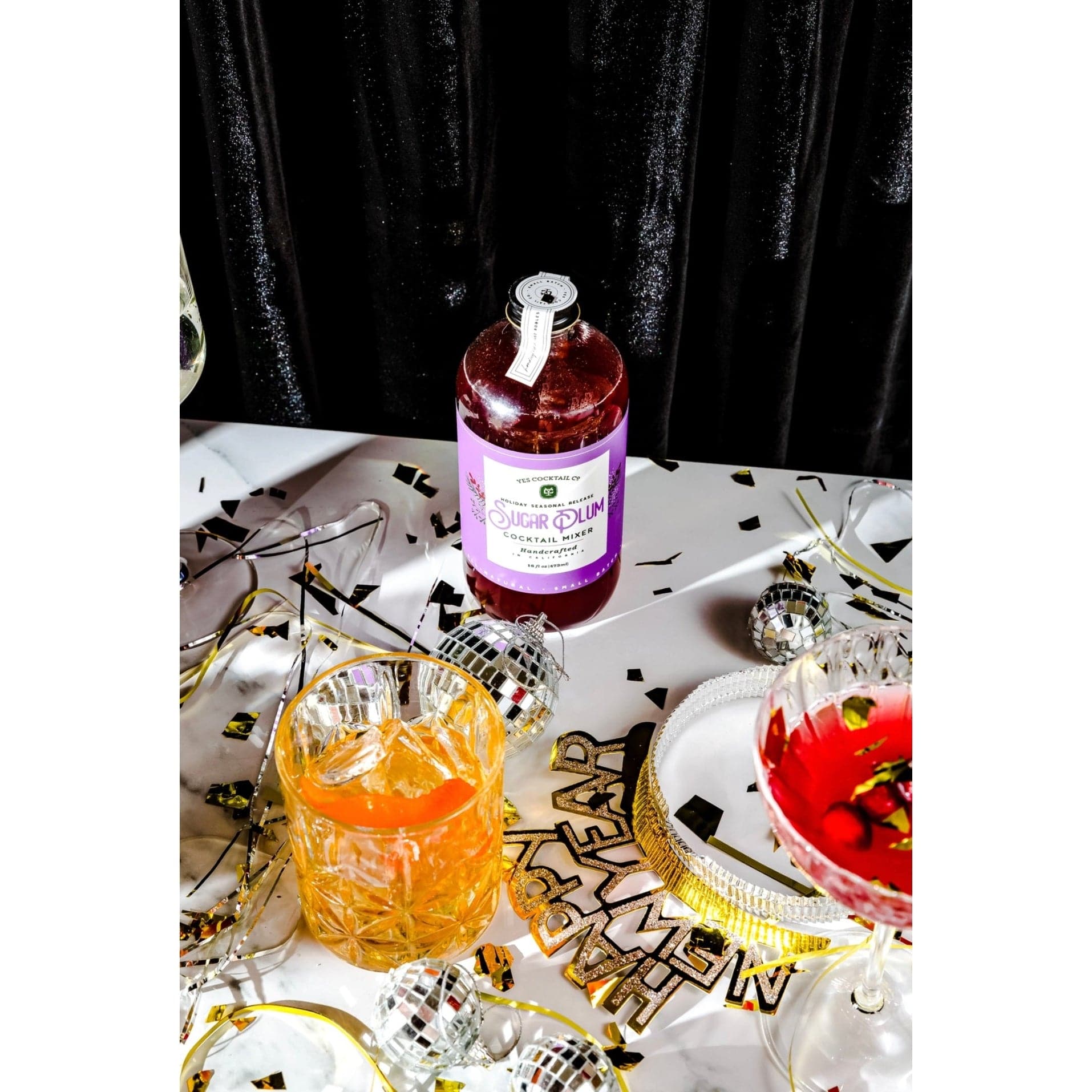 bottle of Sugar Plum Cocktail Mixer displayed on a table decorated for a New Year's Eve party with a finished cocktail