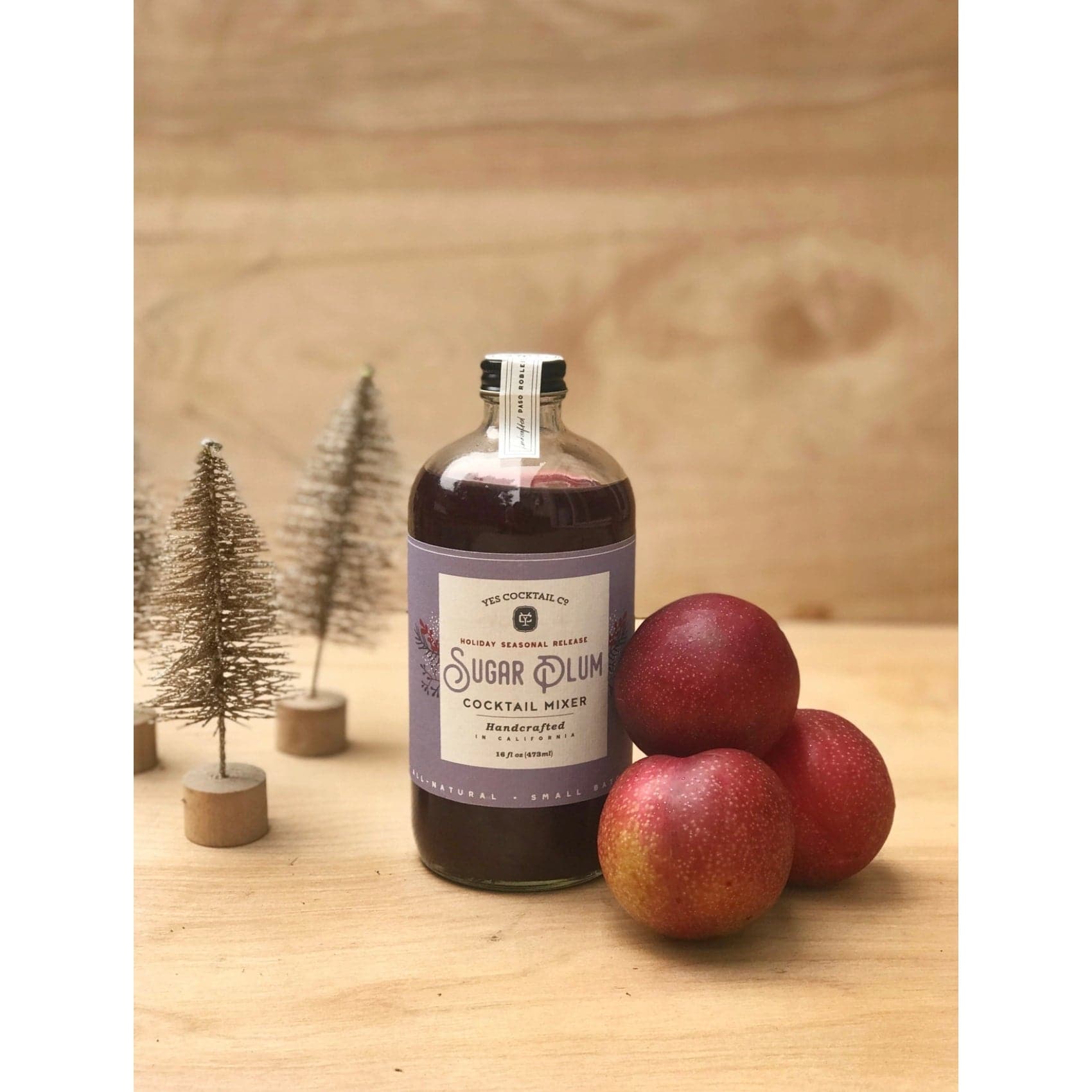 16 oz bottle of hand crafted Sugar Plum Cocktail Mixer made by Yes Cocktail Co on a wood table displayed next to a stack of plums