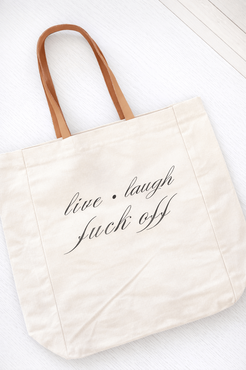 Beige tote bag with 'live laugh fuck off' text on a white background