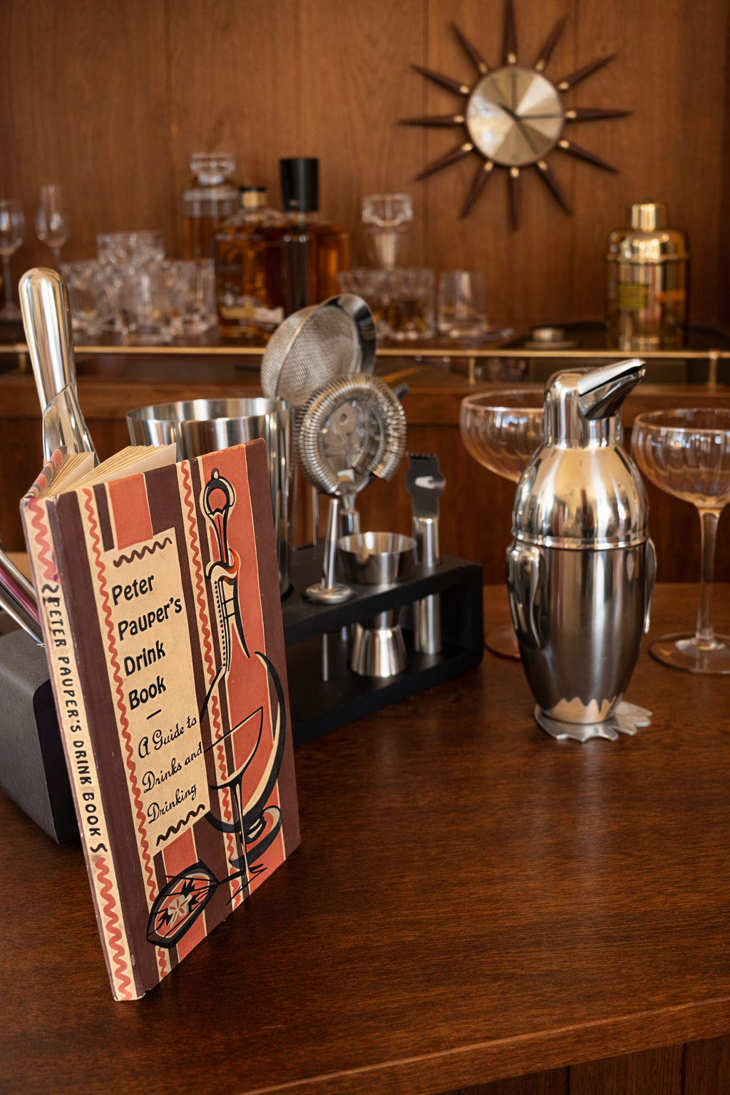 Vintage Peter Pauper’s Drink Book styled with classic stainless steel cocktail shaker and bar tools in a warm midcentury modern home bar setting.