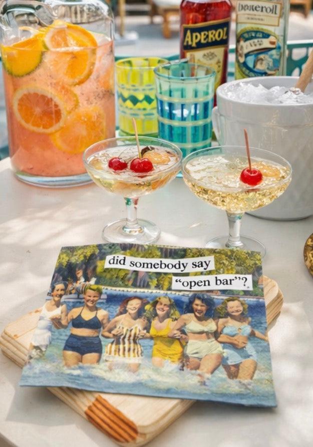 Vintage “Did Somebody Say Open Bar?” cocktail napkins displayed with champagne cocktails, Aperol spritz pitcher, and colorful midcentury modern glasses on a retro cocktail party bar setup.