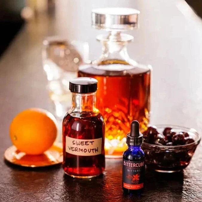 Bitters displayed with a bottle of bourbon, orange, cherries, and vermouth