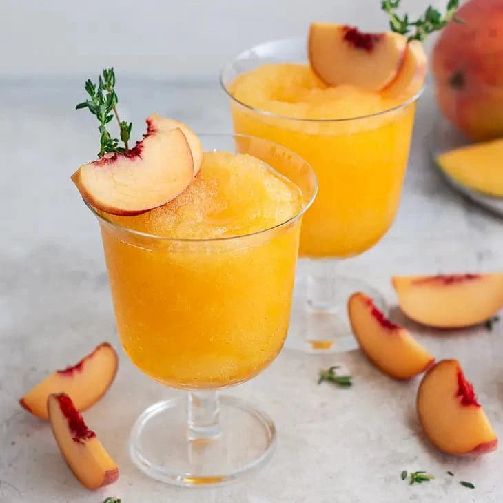 Two glasses of Peach Mango Wine Slushy Mix – Craft Frozen Cocktail Mixer with slices of peaches on a table