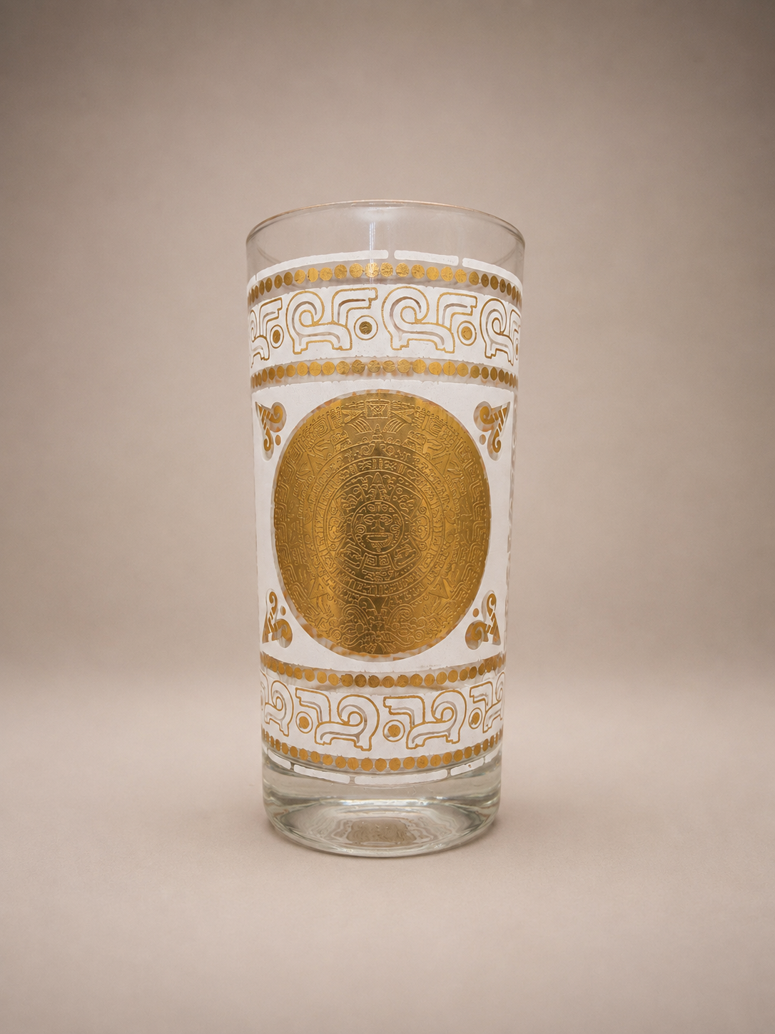 Close-up of a vintage highball glass featuring an intricate gold Aztec-inspired medallion and white geometric pattern.