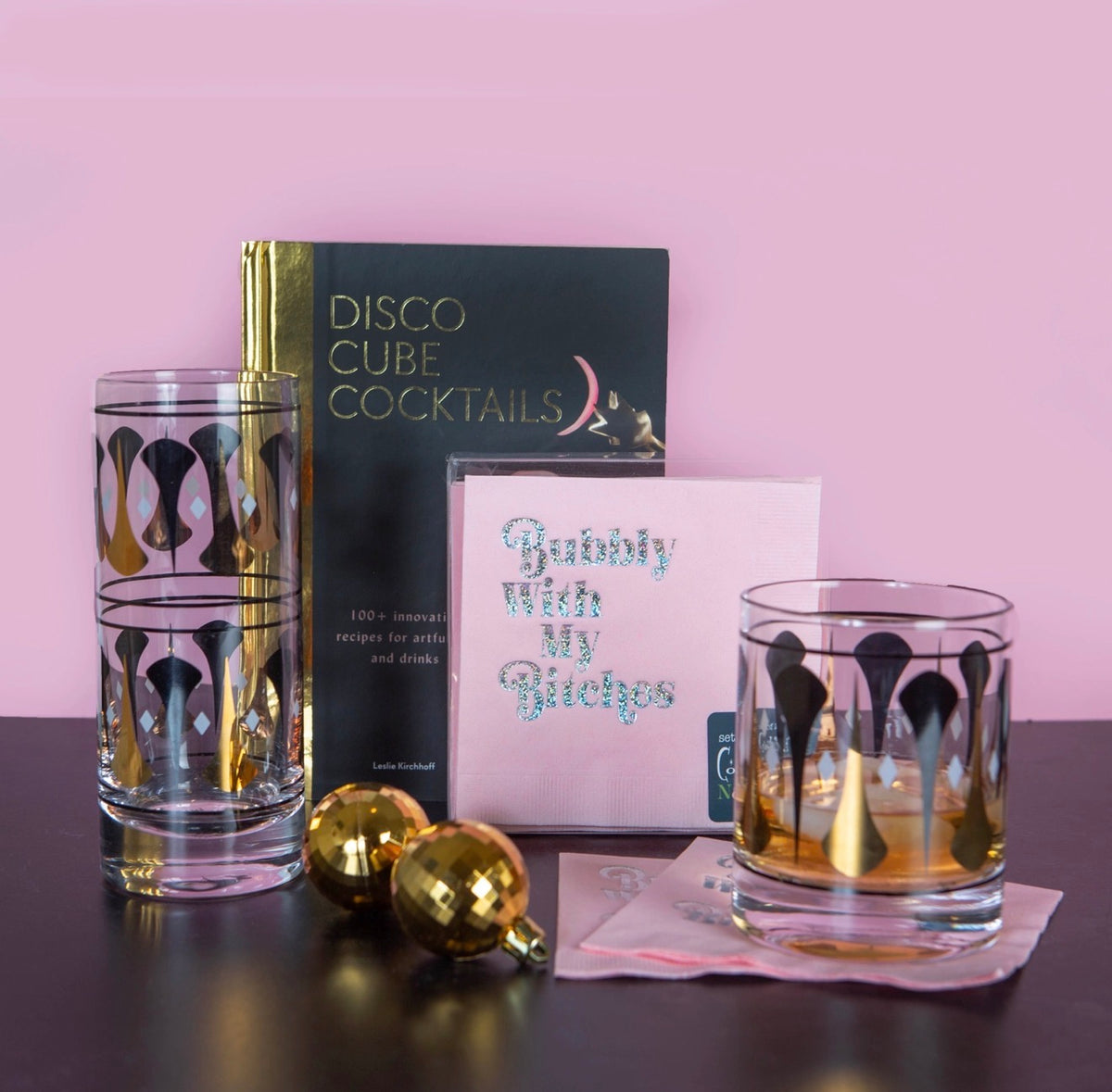 Black and gold whiskey rocks glass styled with retro cocktail books, napkins and gold accents against a pink backdrop