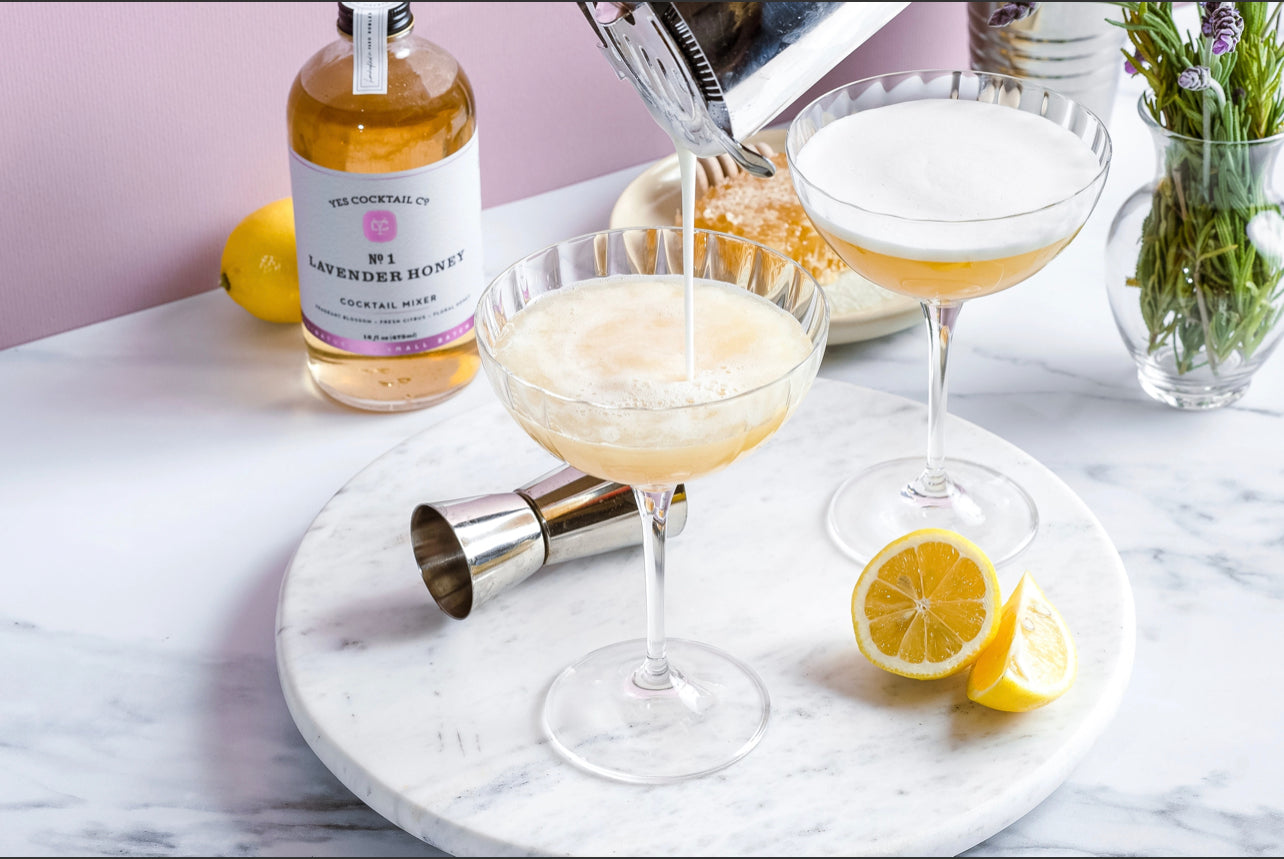 cocktail being poured into two clear martini glasses on a marble countertop with a 16 oz bottle of handcrafted Lavendar and Honey Cocktail Mixer made by Yes Cocktail Co in the background