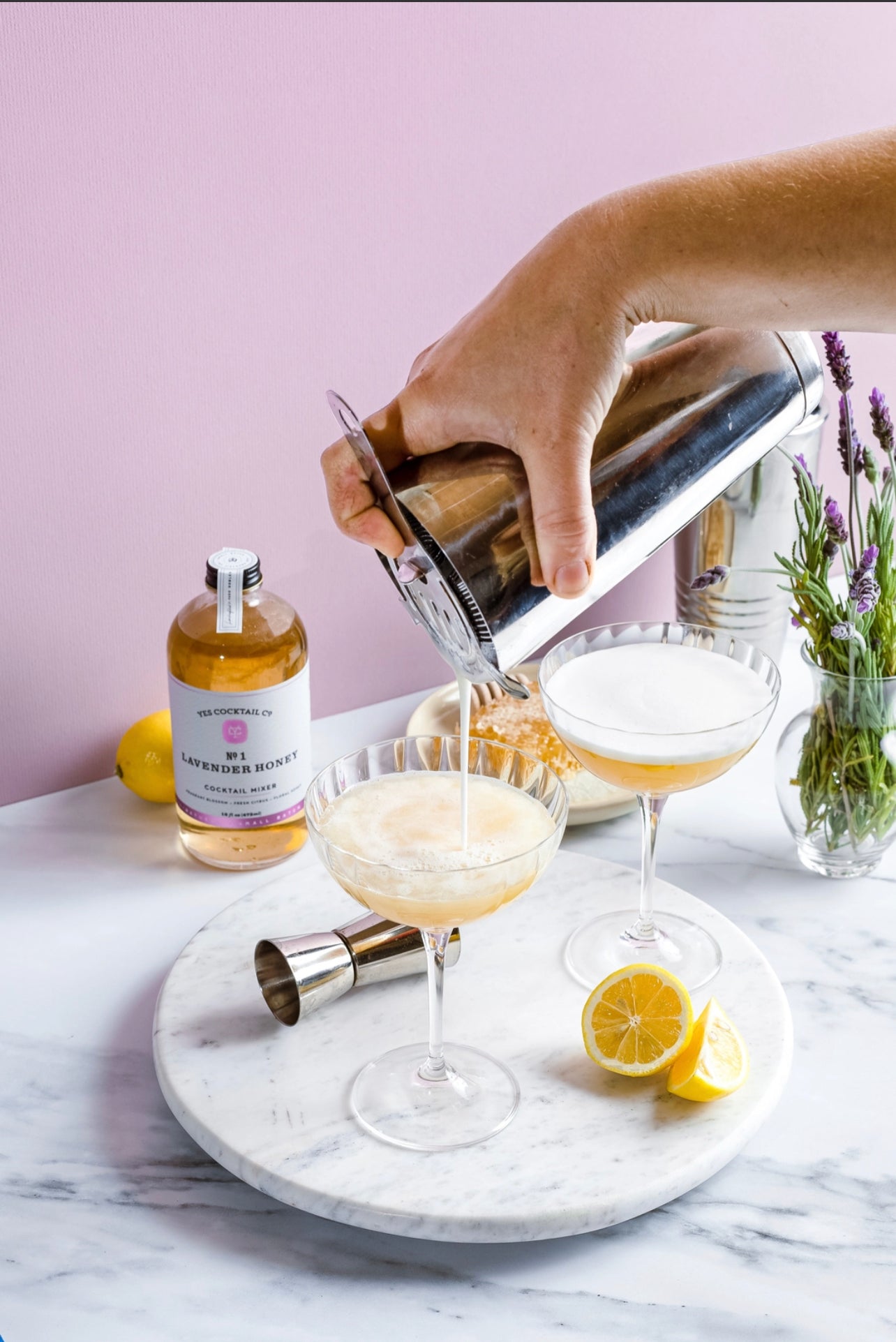 a hand holding a silver cocktail shaker pouring drinks into martinin glasses with the 16 oz bottle of handcrafted Lavendar and Honey Cocktail Mixer made by Yes Cocktail Co in the background