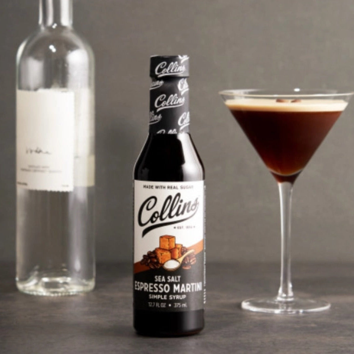 Collins Sea Salt Espresso Martini syrup bottle with a cocktail and a bottle of spirits on a gray background