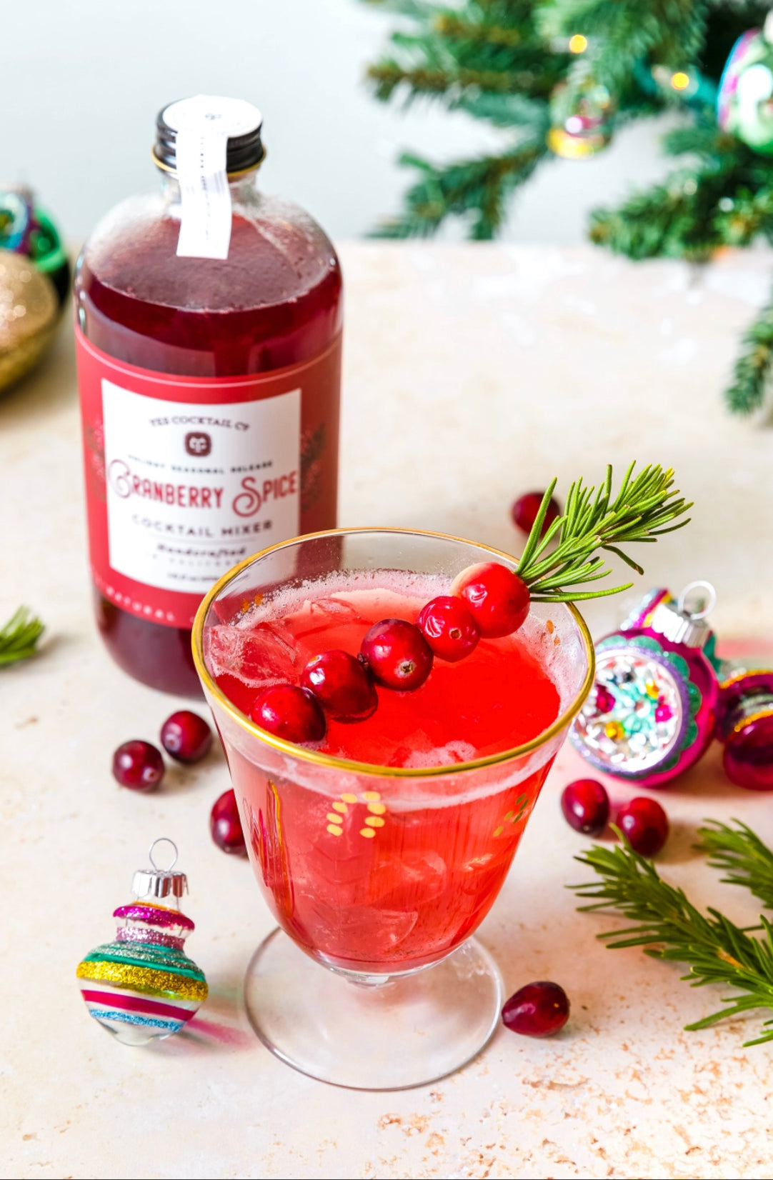 16 oz bottle of handcrafted Cranberry Spice Cocktail Mixer made by Yes Cocktail Co displyed with a finished cocktail in a clear glass garnished with fresh cranberries. Christmas decorations surround the bottle and drink with a tree in the background.