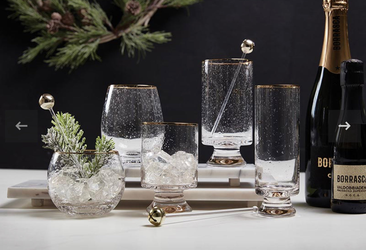 Holiday display of champagne bottles and pine featuring complementing wine glasses, champagne glass, and double old fashioned glass with seeded glass and gold rims