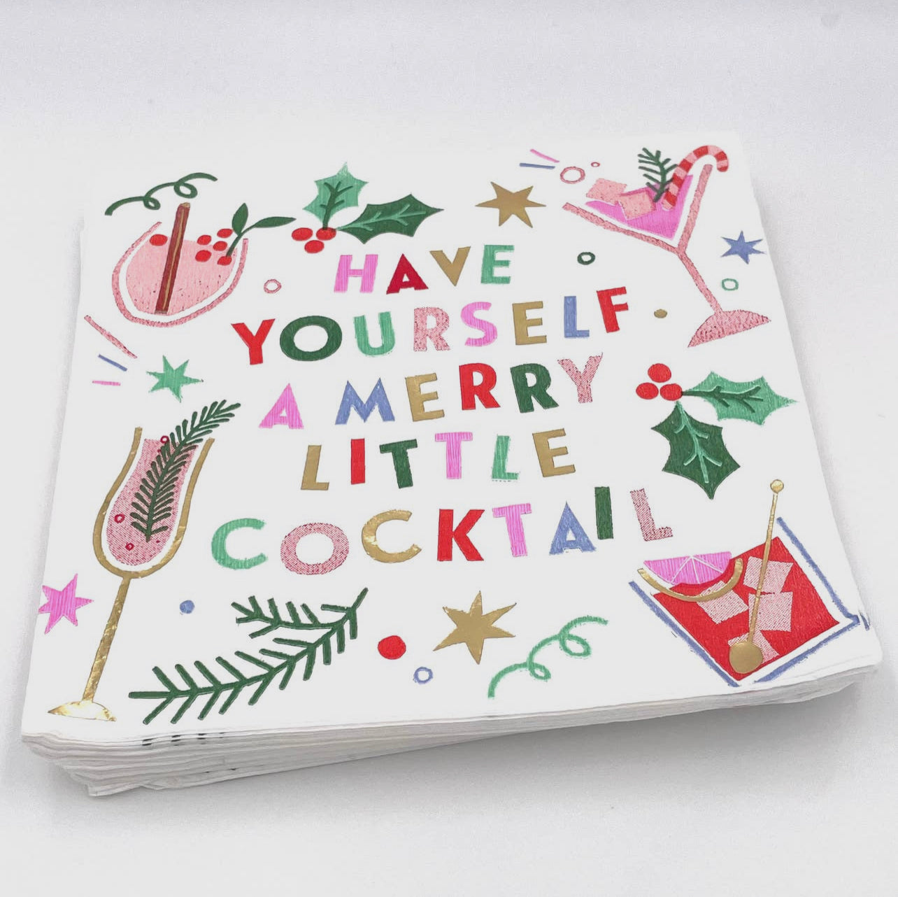 holiday cocktail napkins with a white background featuring different cocktail glasses, holly berries, and stars in festive colors with the words "have yourself a merry little cocktail" in the center and bits of gold foil for accent