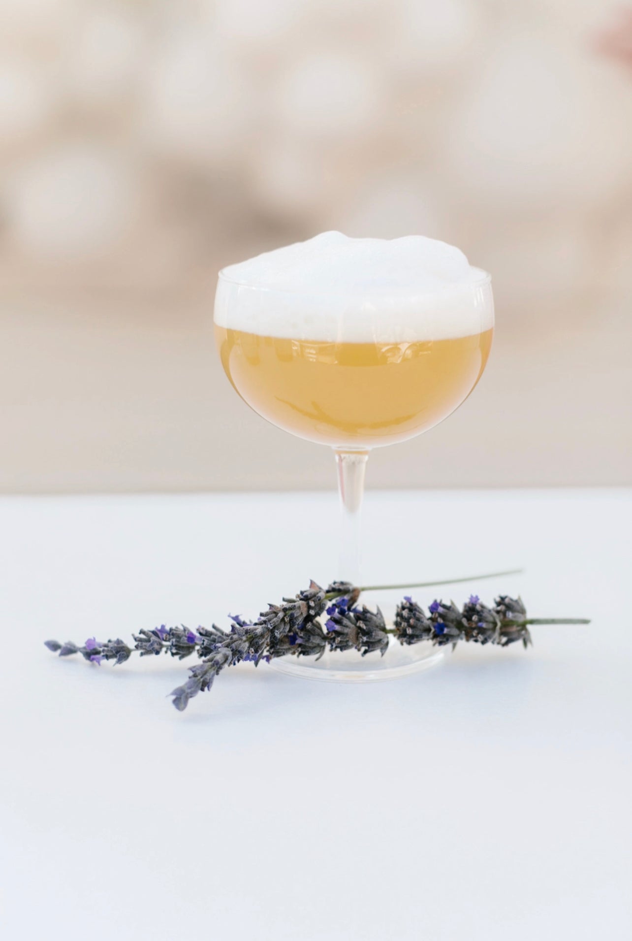 a golden cocktail in a clear martini glass with a soft foam top accented with fresh lavender