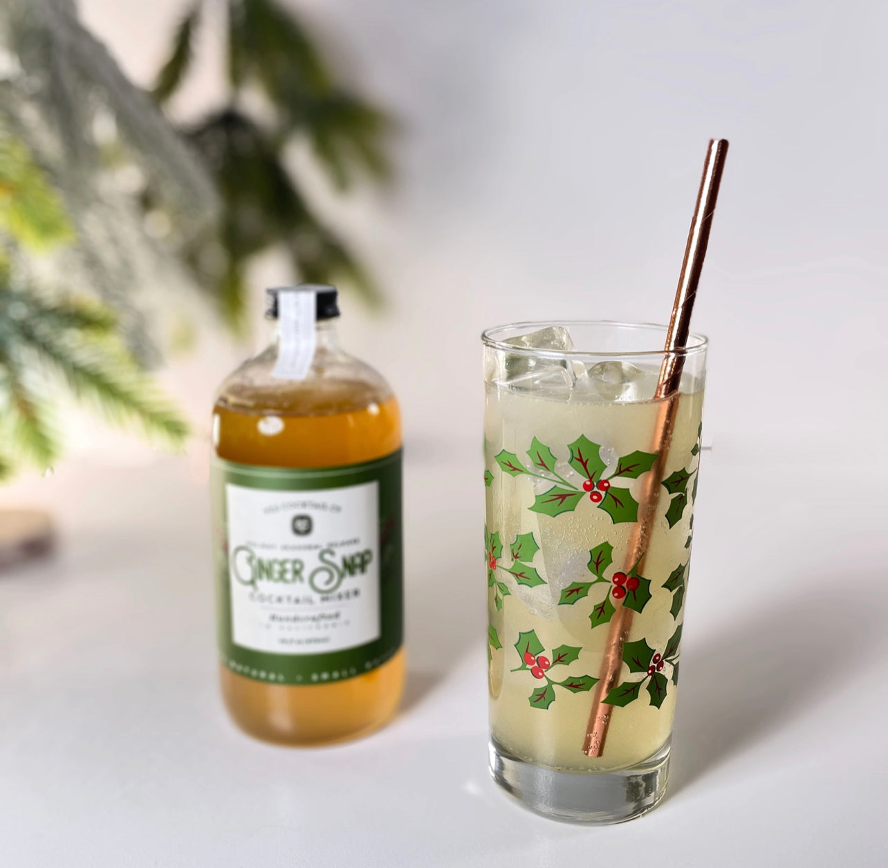 16 oz bottle of handcrafted Ginger Snap Cocktail Mixer made by Yes Cocktail Co displayed with a finished cocktail in a holiday decorated glass with stainless steel straw