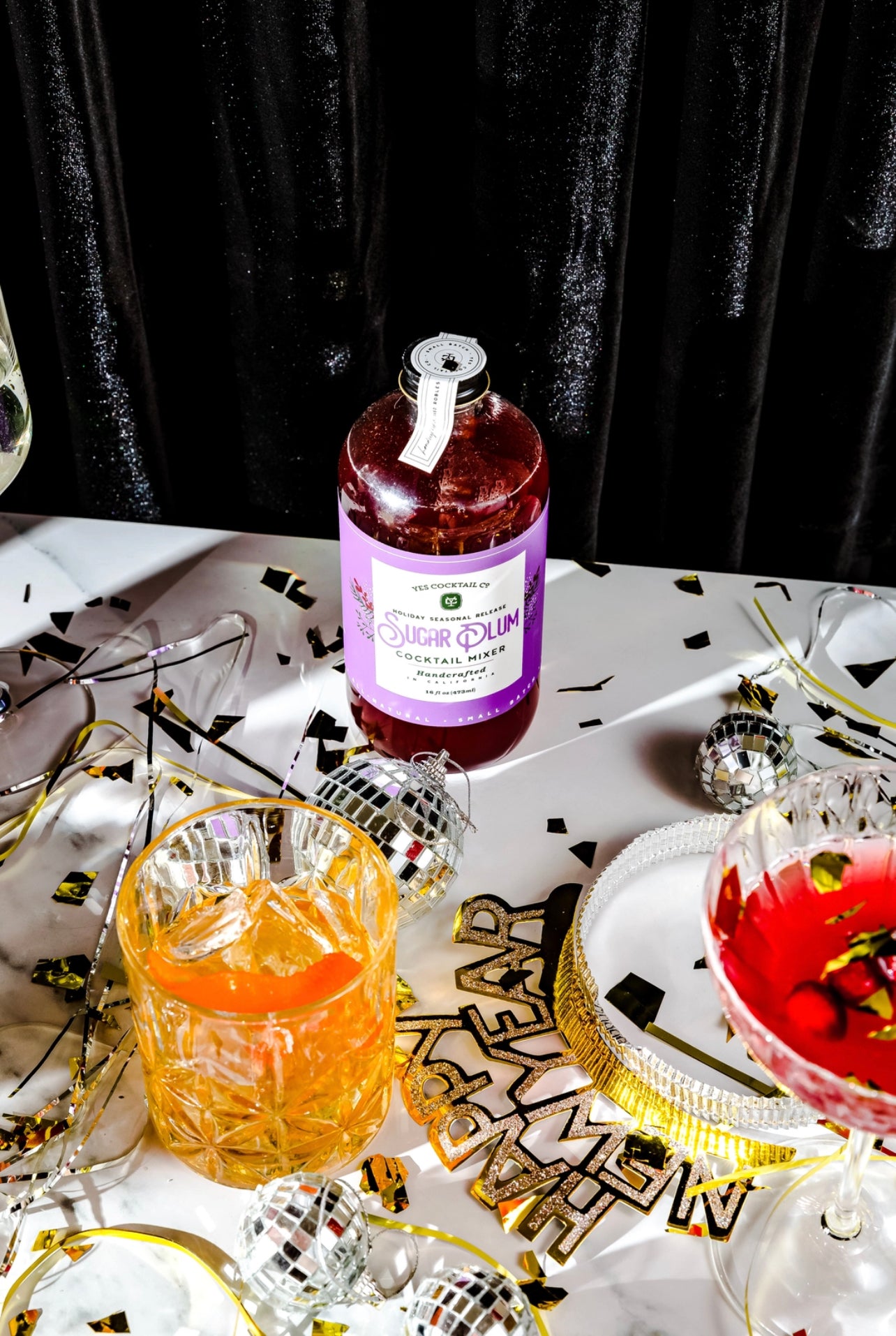 bottle of Sugar Plum Cocktail Mixer displayed on a table decorated for a New Year's Eve party with a finished cocktail