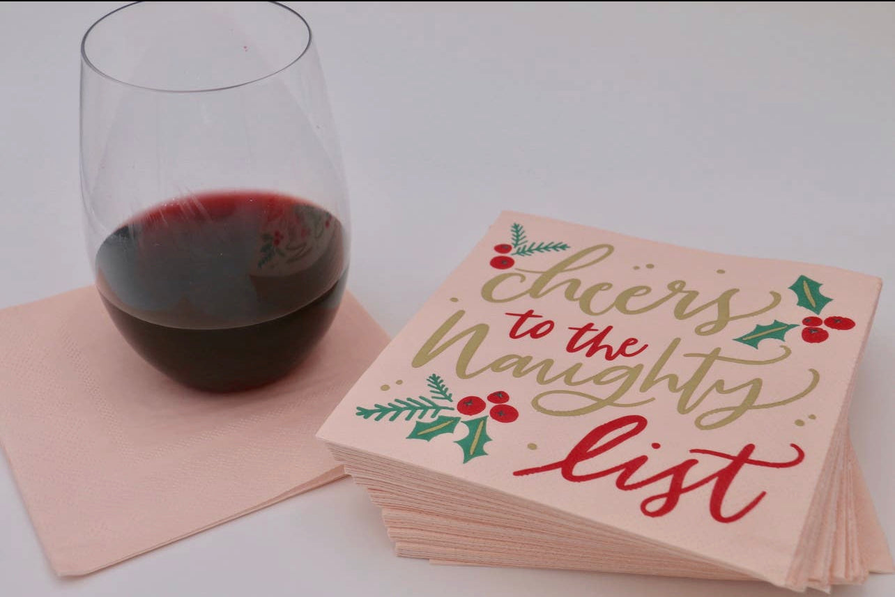 Christmas cocktail napkins with a pink background decorated with holly berries and the words "cheers to the naughty list" in a fancy script, in red and gold, displayed with a wine glass