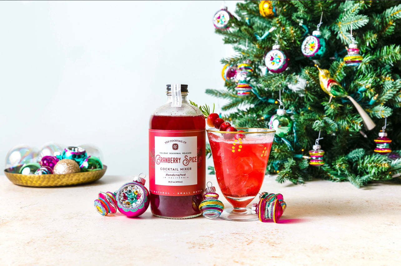 16 oz bottle of handcrafted Cranberry Spice Cocktail Mixer made by Yes Cocktail Co displyed with a finished cocktail in a clear glass garnished with fresh cranberries. Christmas decorations surround the bottle and drink with a tree in the background.