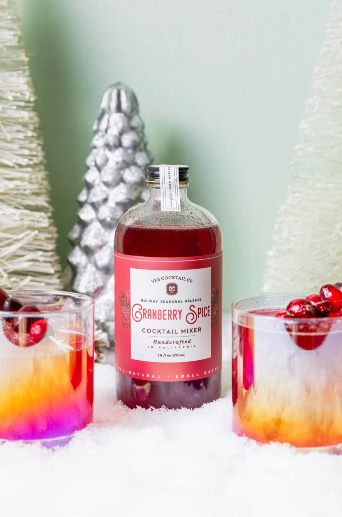 16 oz bottle of handcrafted Cranberry Spice Cocktail Mixer made by Yes Cocktail Co displayed with finished cocktails in iridescent double old fashioned glasses that are garnished with fresh cranberries. Fake snow is spread on the table with decorative Christmas tree shaped decorations in the background.