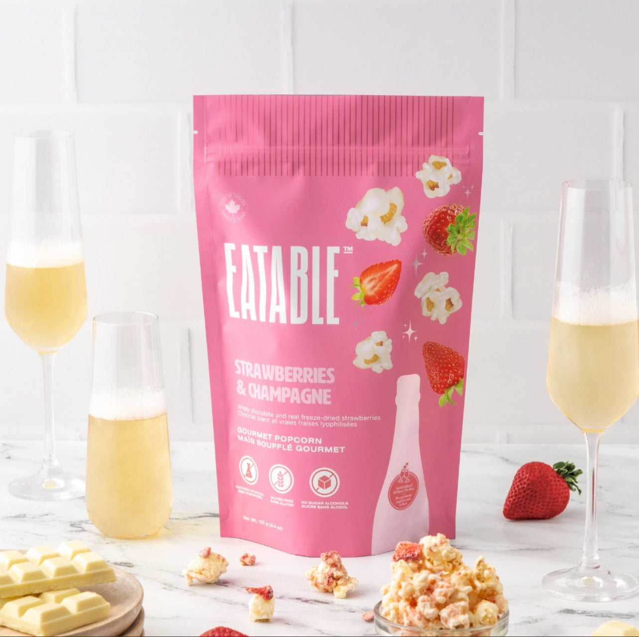 Pink package of 'Eatable' strawberries and champagne popcorn on a white surface with glasses of champagne and strawberries.