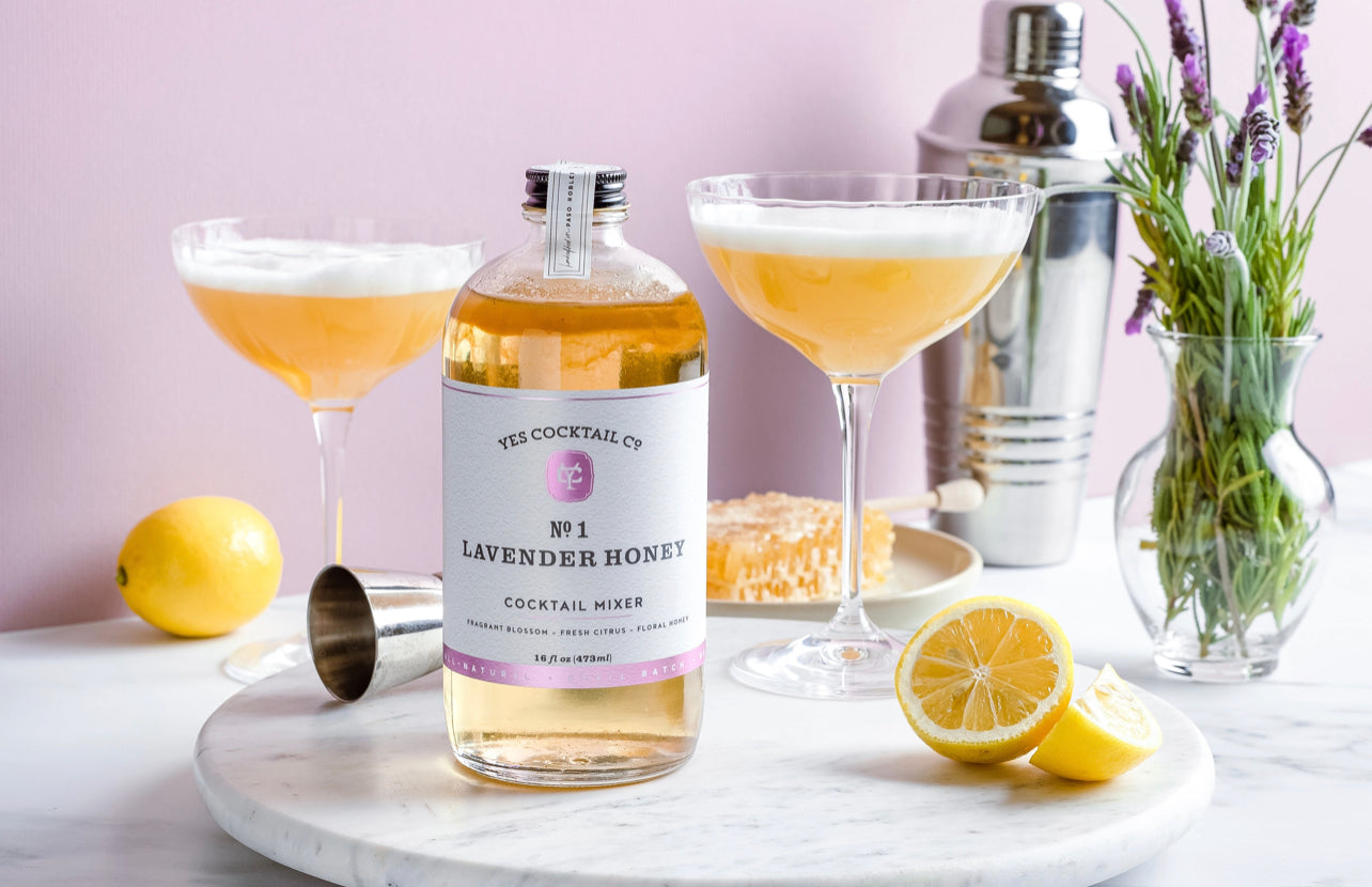 16 oz bottle of handcrafted Lavendar and Honey Cocktail Mixer made by Yes Cocktail Co displayed on marble with cocktail filled martini glasses flanking each side and fresh lemons and lavender for accent