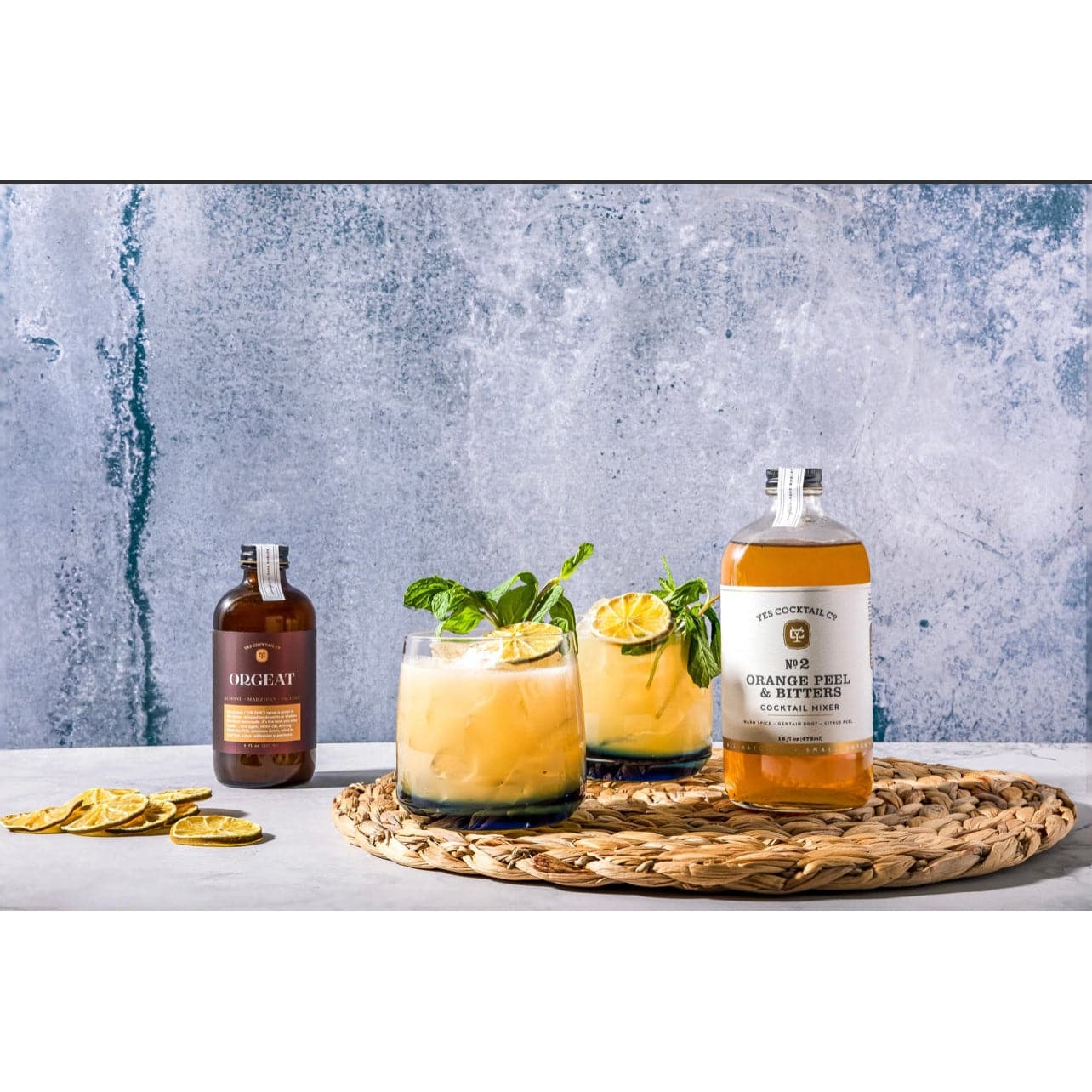 display of the Orange Peel and Bitters Cocktail Mixer on a marble countertop surrounded by  dehydrated fruit, a bottle of orgeat syrup, and cocktails