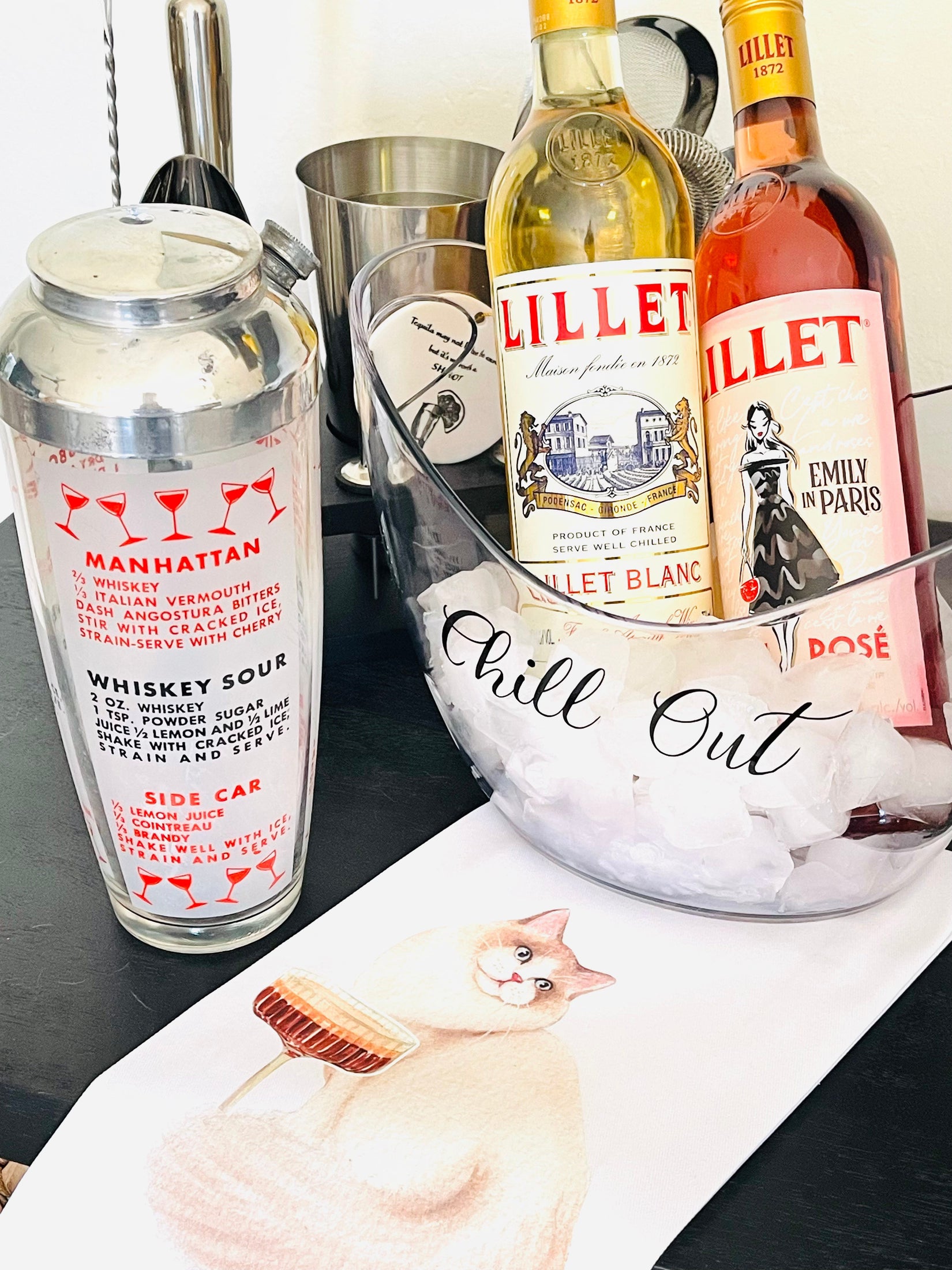 Two bottles of Lillet with a cocktail shaker and ice bucket on a table.