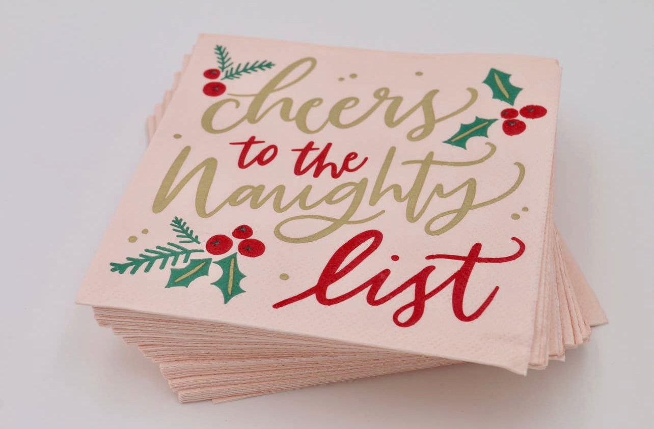 Christmas cocktail napkins with a pink background decorated with holly berries and the words "cheers to the naughty list" in a fancy script, in red and gold,