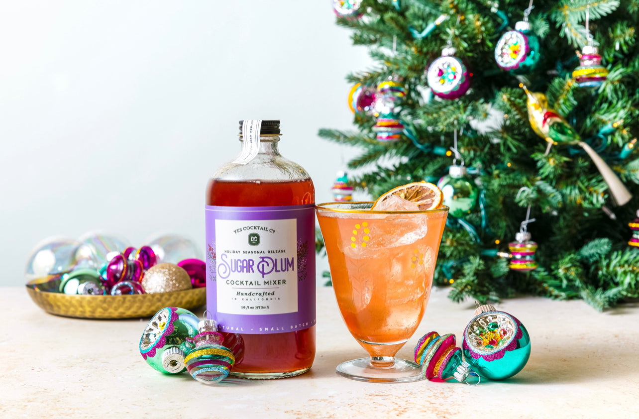16 oz bottle of hand crafted Sugar Plum Cocktail Mixer made by Yes Cocktail Co displayed next to a cocktail made with the mix surrounded by Christmas ornaments and a tree in the background