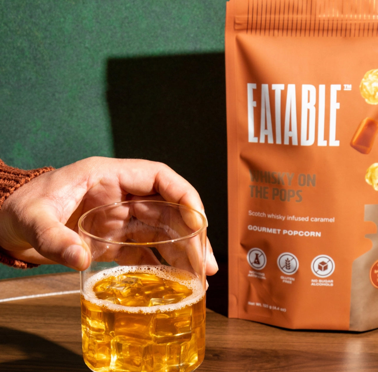 Hand holding a glass of amber liquid next to an 'Eatable' product package on a wooden surface.