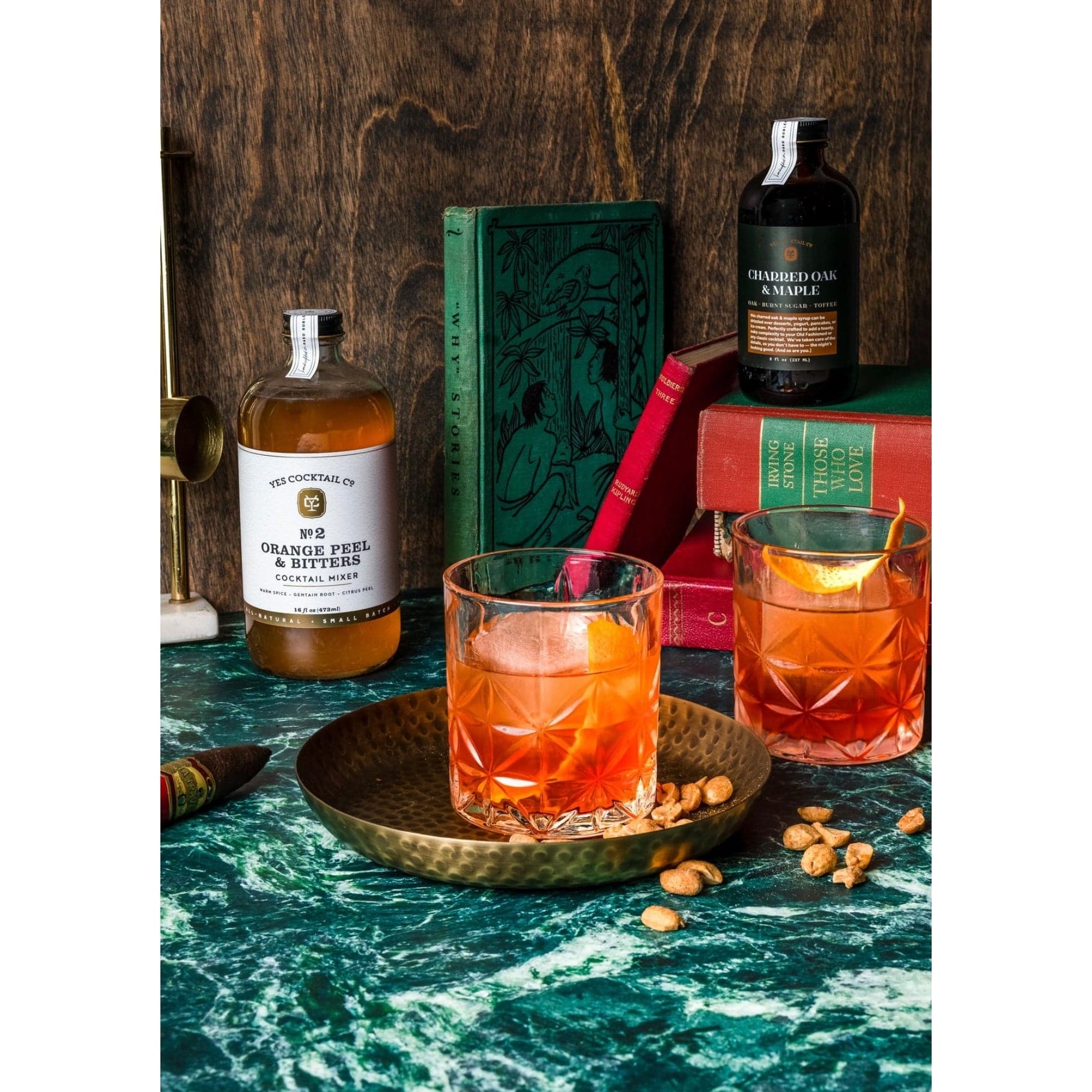 display of the Orange Peel and Bitters Cocktail Mixer on a marble countertop surrounded by  cocktails in double old fashioned glasses in a den setting with books