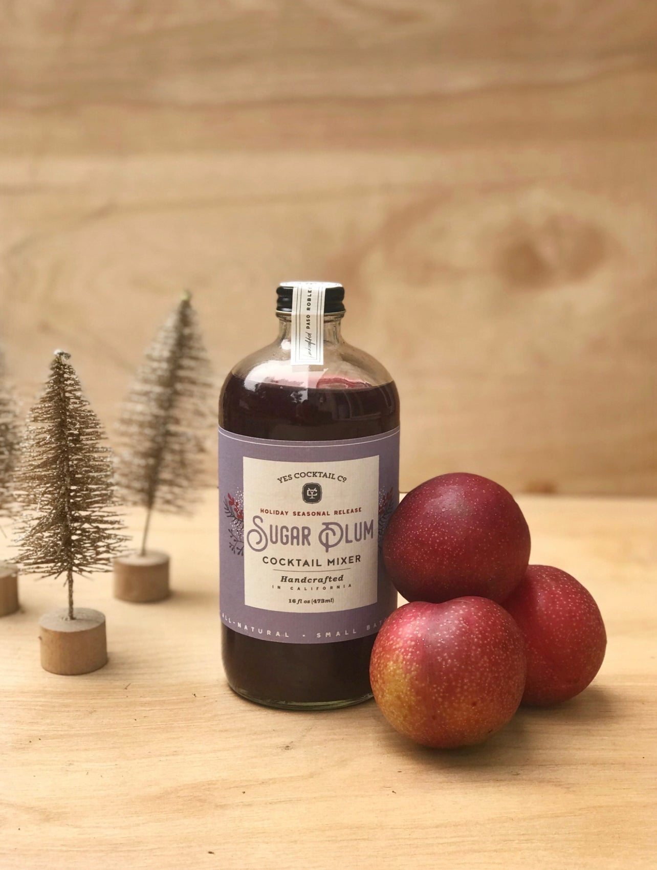 16 oz bottle of hand crafted Sugar Plum Cocktail Mixer made by Yes Cocktail Co on a wood table displayed next to a stack of plums