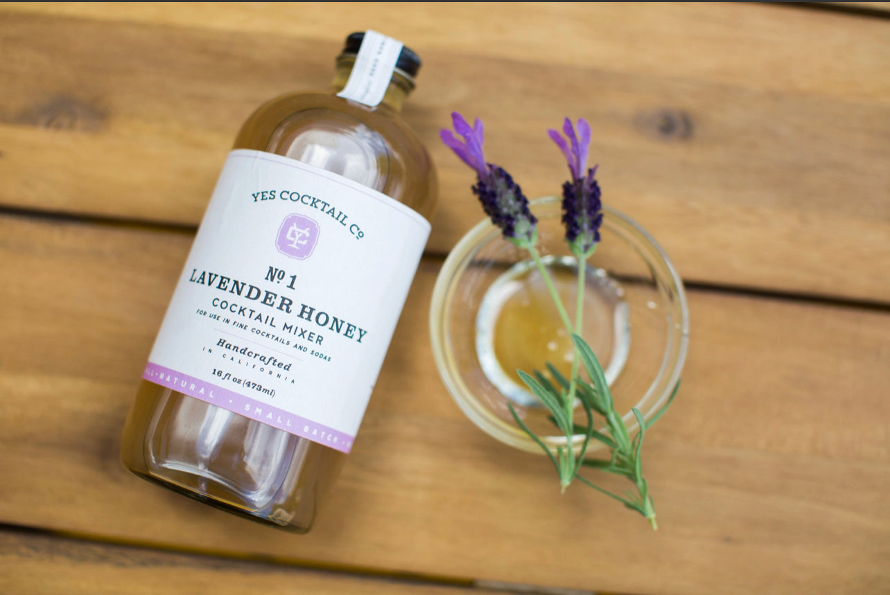 16 oz bottle of handcrafted Lavendar and Honey Cocktail Mixer made by Yes Cocktail Co laying on a wooden surface  next to a small dish of honey with lavender laying across the top