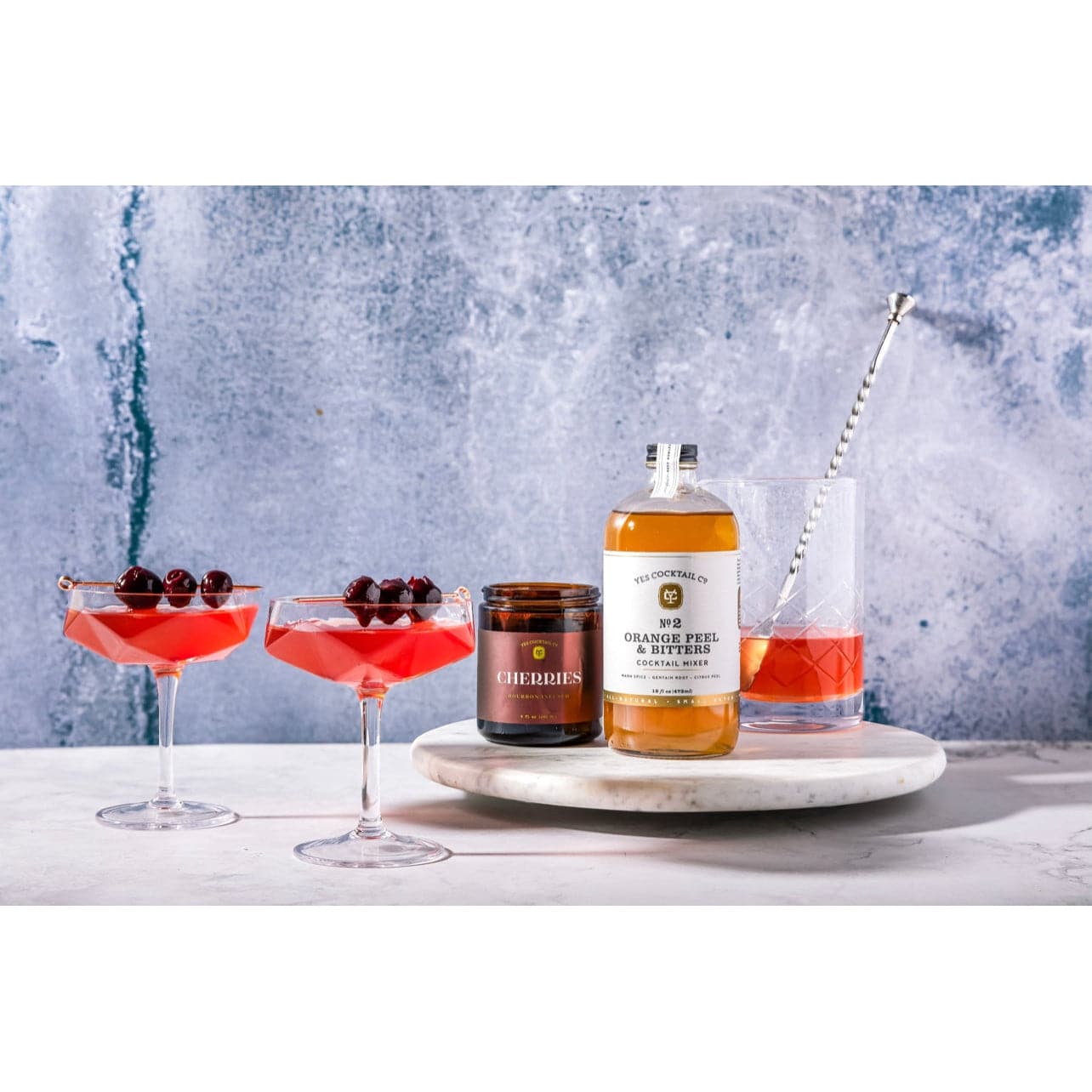 display of the Orange Peel and Bitters Cocktail Mixer on a marble countertop surrounded by  cocktails in martini glasses garnished with bourbon cherries