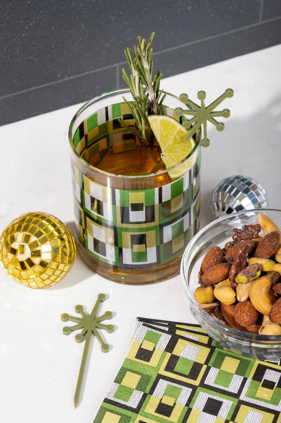 Green geometric rocks cocktail glass styled with a whiskey cocktail, citrus garnish and retro atomic drink picks