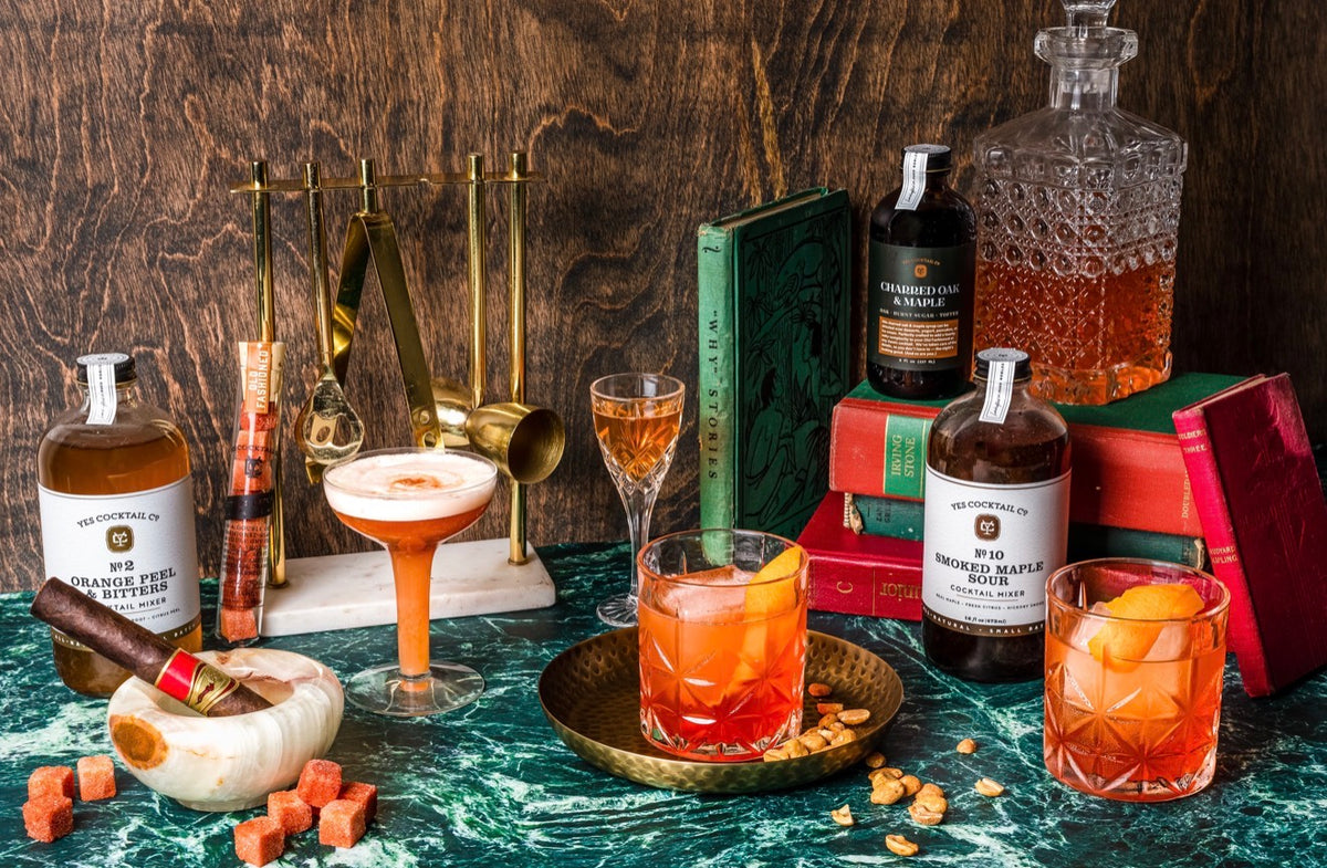Vintage-inspired cocktail bar with whiskey drinks, orange-garnished Old Fashioned cocktails, brass bar tools, a crystal decanter and Yes Cocktail Co. mixers.