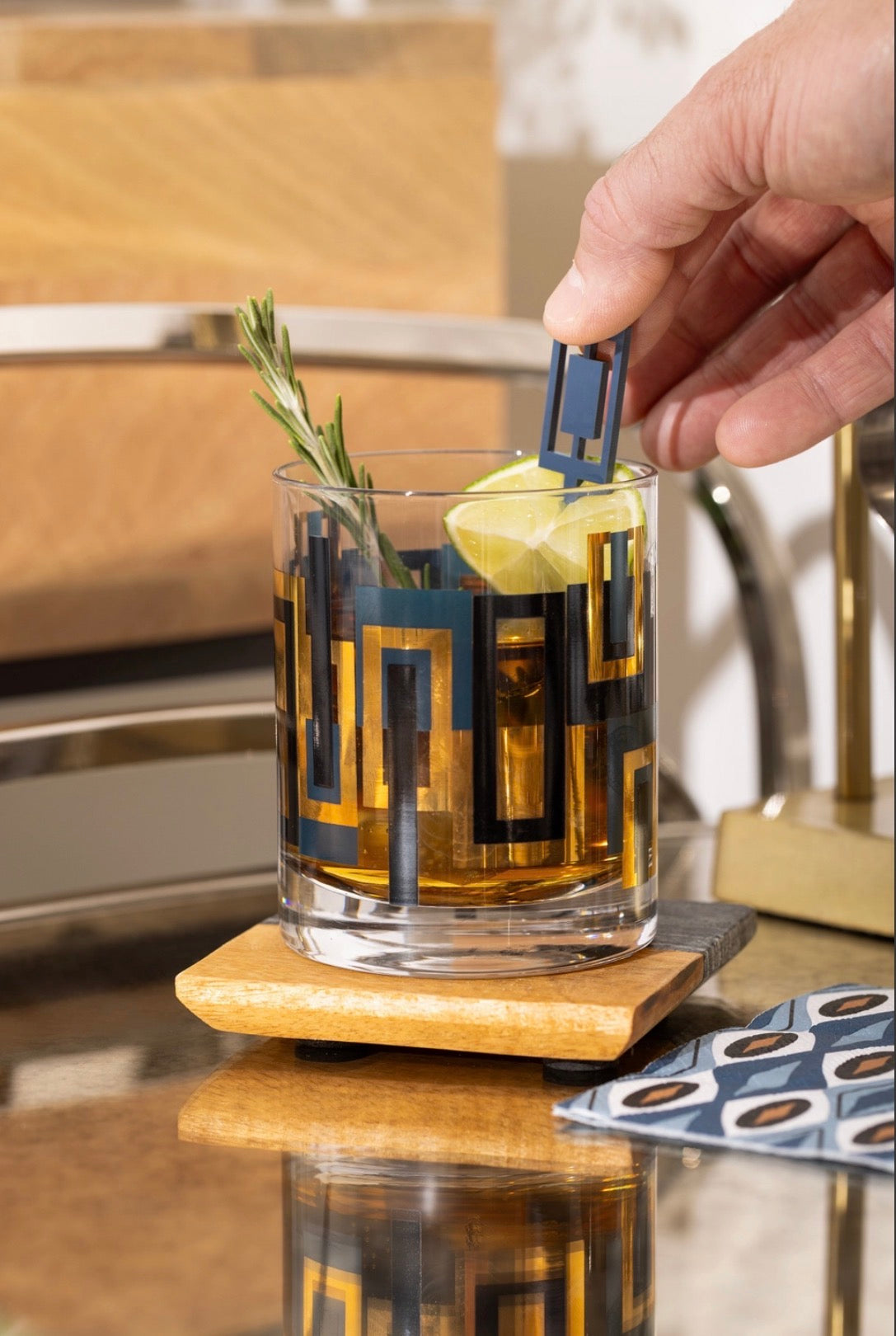 Blue, black and gold geometric cocktail rocks glass styled with whiskey, lime and rosemary on a retro bar
