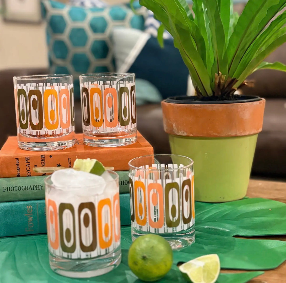Four mid century inspired cocktail glasses with colorful oval geometric patterns styled with vintage books, tropical greenery and limes