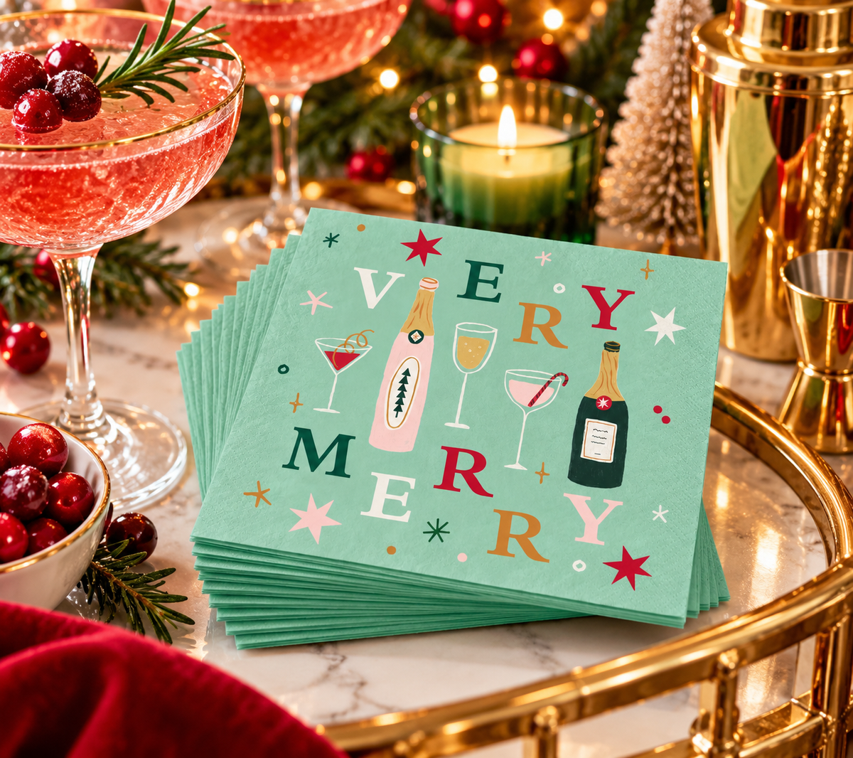 Very Merry holiday cocktail napkins styled on a festive Christmas bar with pink cocktails, cranberries, greenery and gold barware.