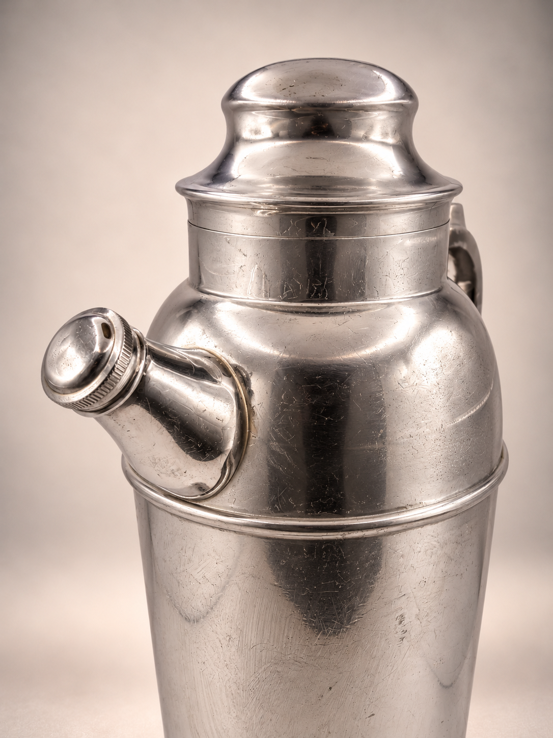 Close-up of vintage metal cocktail shaker showing angled capped pour spout, rounded lid, and scratches across the silver-tone finish.