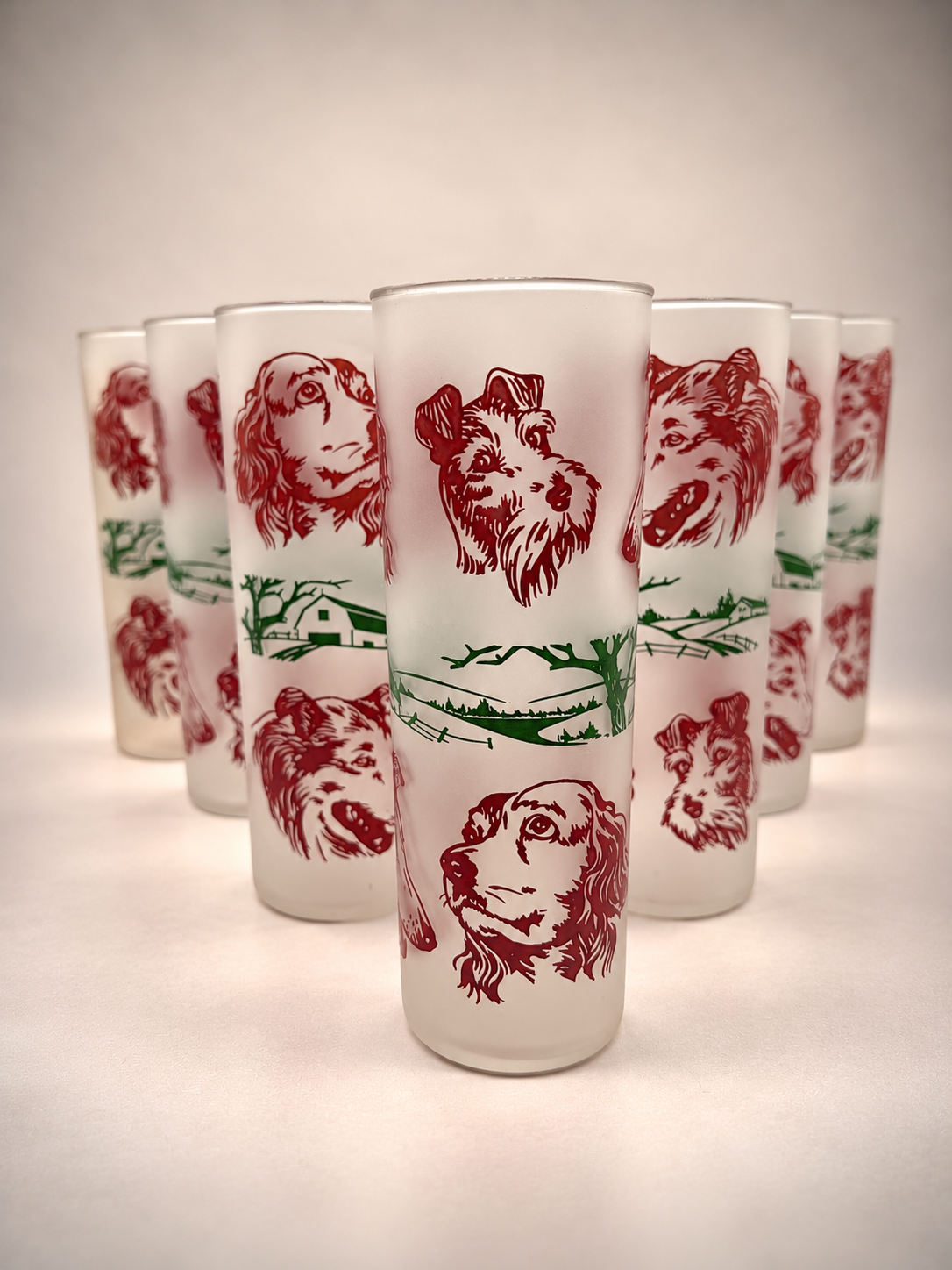 Seven vintage frosted dog portrait highball glasses arranged together, featuring cranberry-red canine illustrations and green countryside designs.