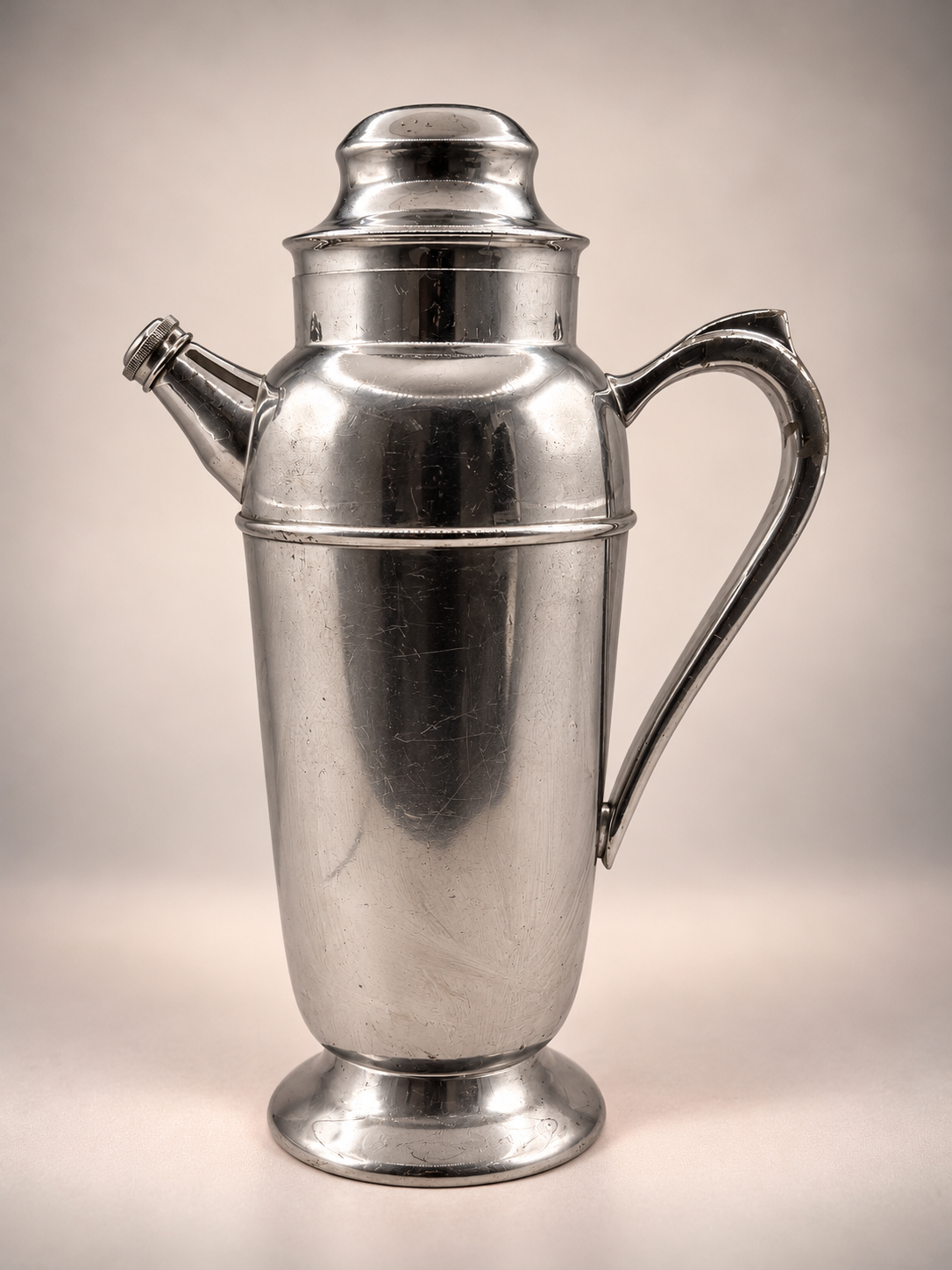 Full front view of vintage silver-tone cocktail shaker featuring curved handle, angled pour spout, rounded lid, and pedestal base.