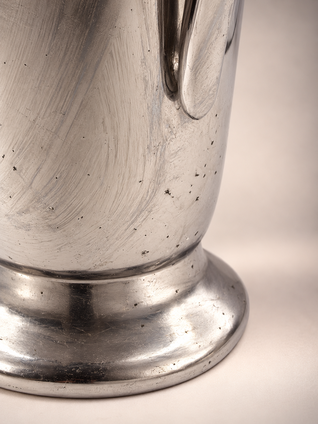 Close-up of silver-tone cocktail shaker lower body and pedestal base showing surface scratches, dark spotting, and age-related finish wear.