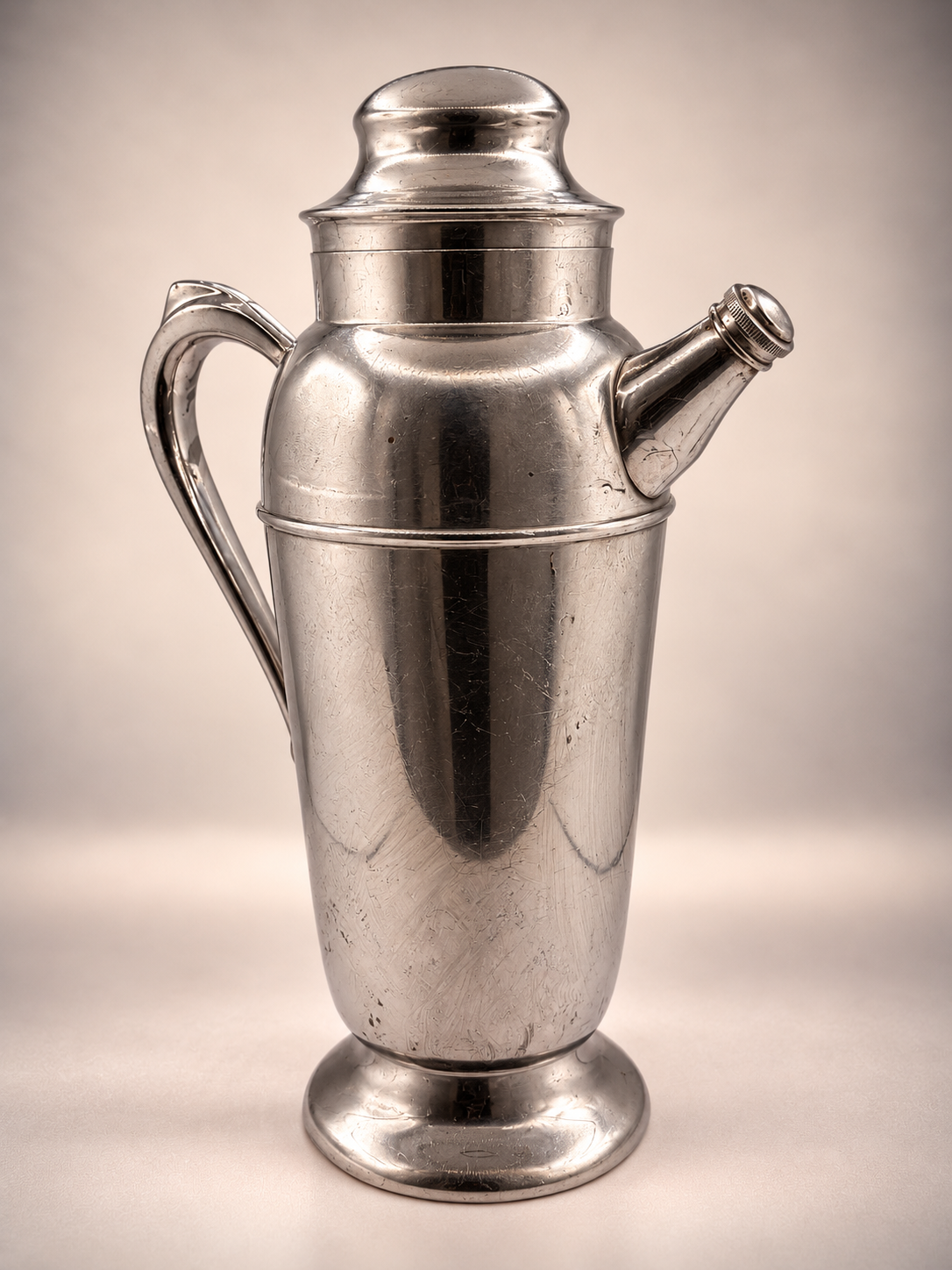 Reverse view of vintage silver-tone cocktail shaker showing its curved handle, angled pour spout, pedestal base, and surface wear.