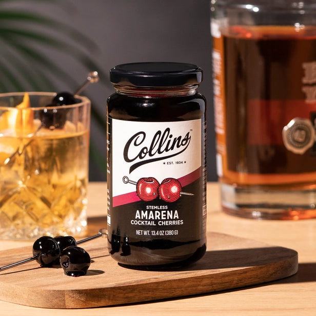 jar of stemless amarena cocktail cherries made by Collins displayed on a wood surface with a bottle and glass of bourbon and cherries on a cocktail pick