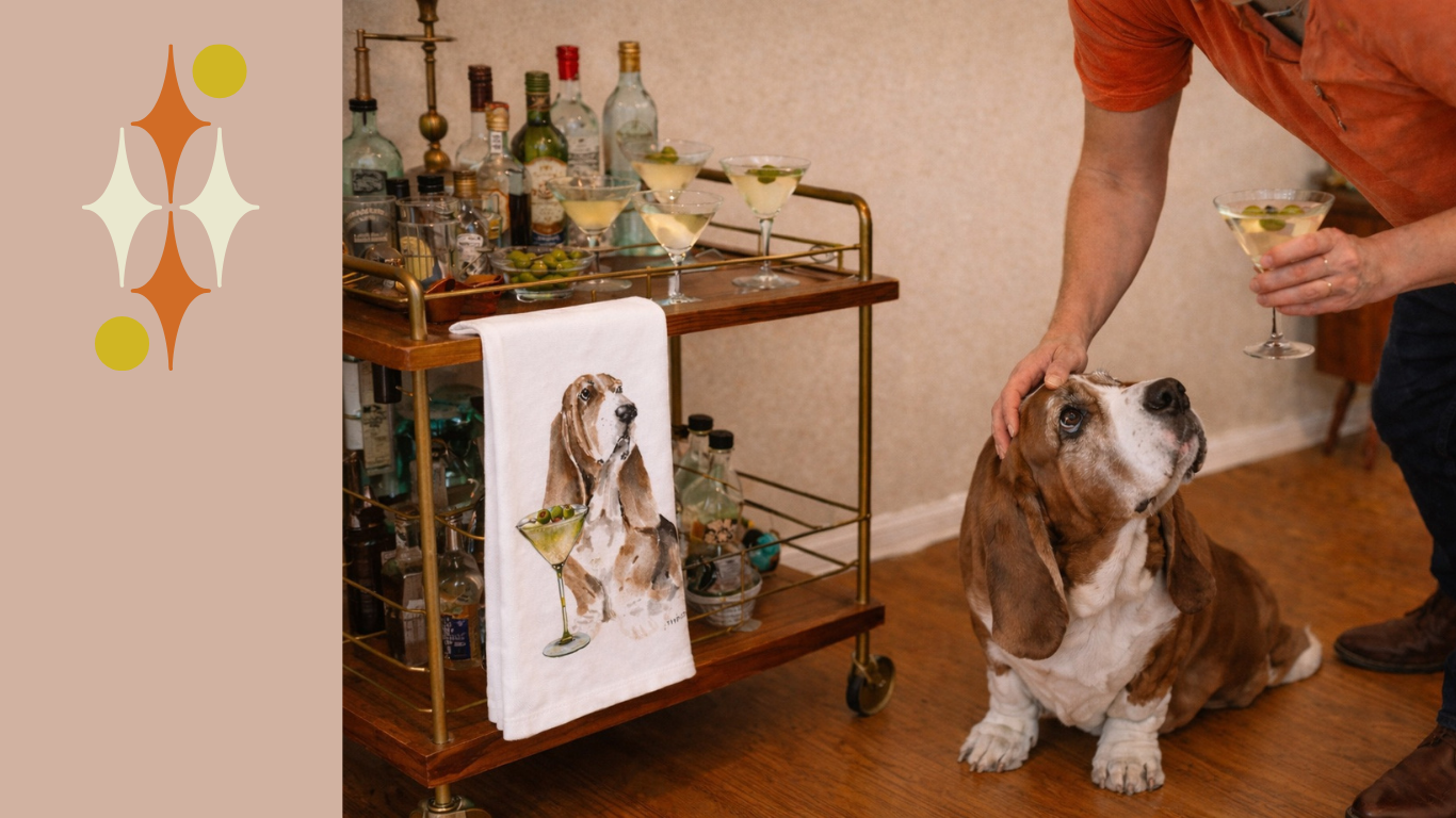 Basset hound sitting beside a vintage bar cart with martini glasses and a dog-themed cocktail towel