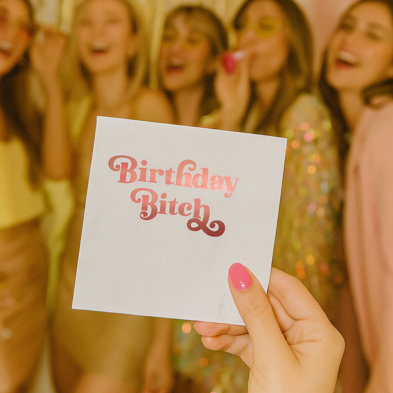 Birthday Bitch cocktail napkin held at birthday party with guests celebrating in background.